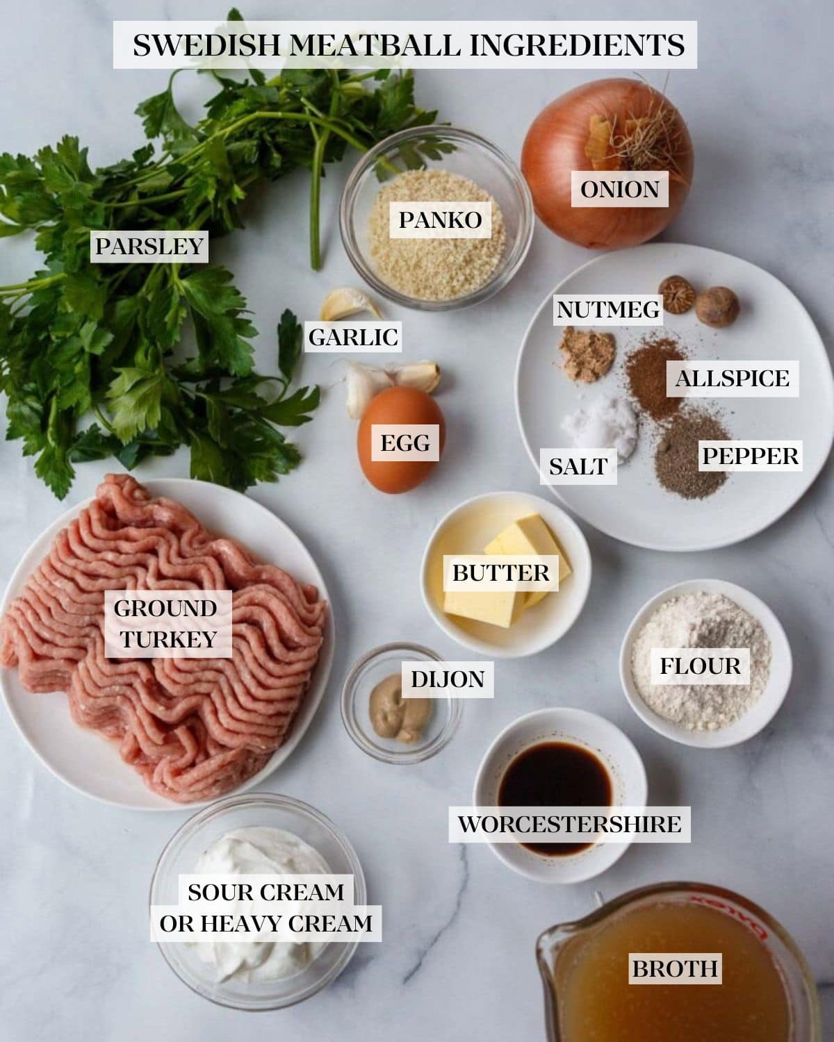 ingredients for Swedish meatballs laid out on marble countertop - parsley bunch, bowl of breadcrumbs, yellow onion, garlic cloves, egg, plate of spices and fresh nutmeg, small bowl of butter cubes, small bowl of flour, tiny dish of Dijon mustard, small bowl of Worcestershire, small bowl of Greek yogurt, and plate of ground turkey.