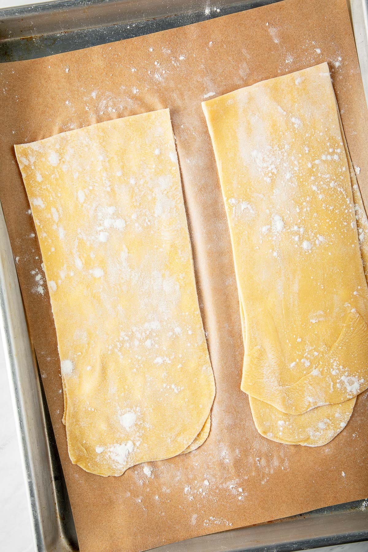 thin sheets of homemade pasta dough folded over on parchment-lined baking sheet with dusting of flour.