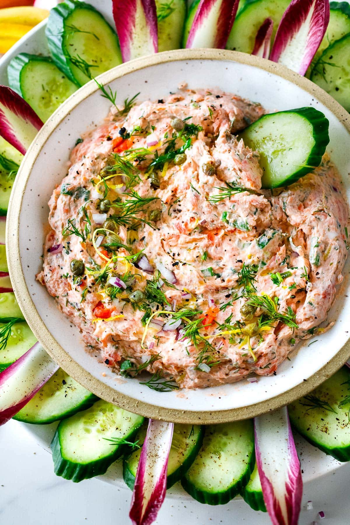 bowl of smoked salmon dip garnished with capers, lemon zest, dill, red onion, with cucumber slice for dipping. 