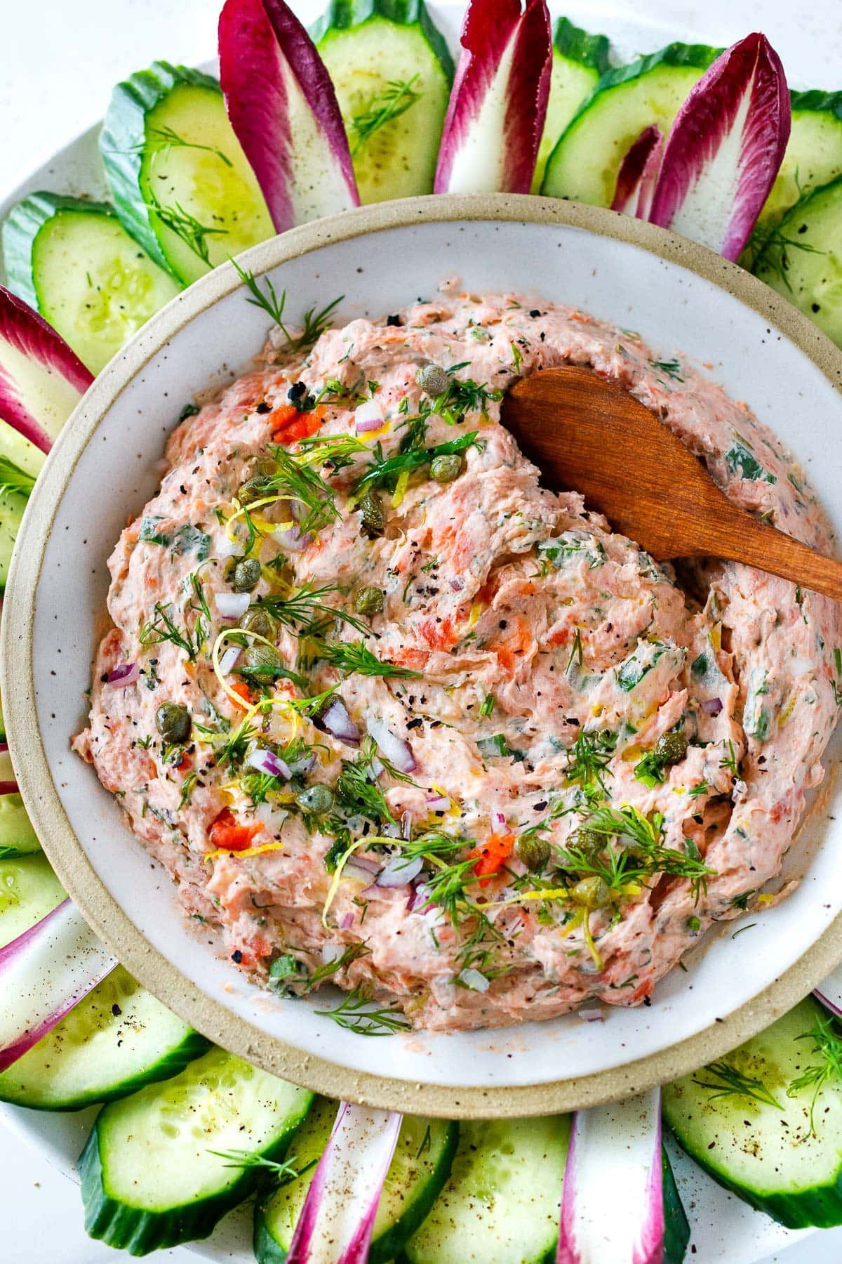 smoked salmon dip in serving bowl surrounded by crudité, garnished with dill, lemon zest, capers, red onion, with wood spoon.