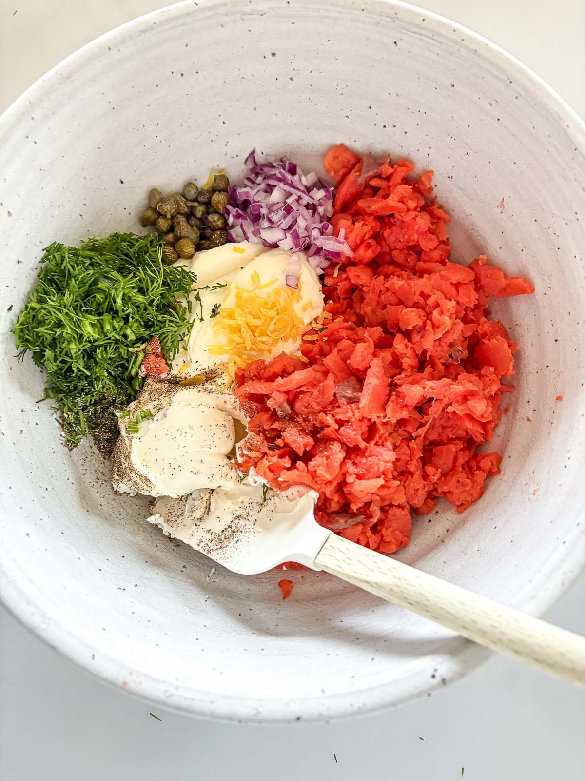 mixing bowl with diced smoked salmon, greek yogurt, cream cheese, lemon zest, red onion, capers, dill, black pepper with spatula. 