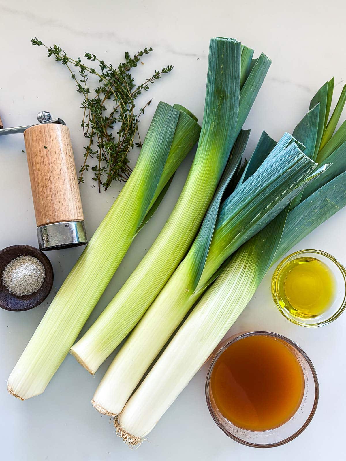 ingredients for braised leeks laid out - thyme sprigs, pepper shaker, flaky salt, four leeks, bowl of oil, bowl of broth.