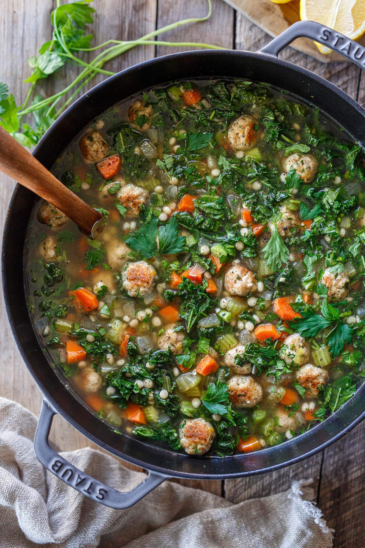 Italian wedding soup in Dutch oven with utensil with meatballs, kale, carrots, onion, pearl couscous, parsley, and broth. 