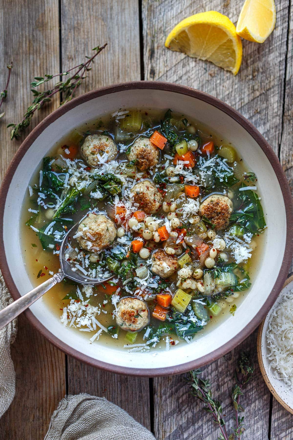 bowl of Italian wedding soup with spoon with seared meatballs, pearl couscous, kale, carrots, onion, celery, parmesan. 