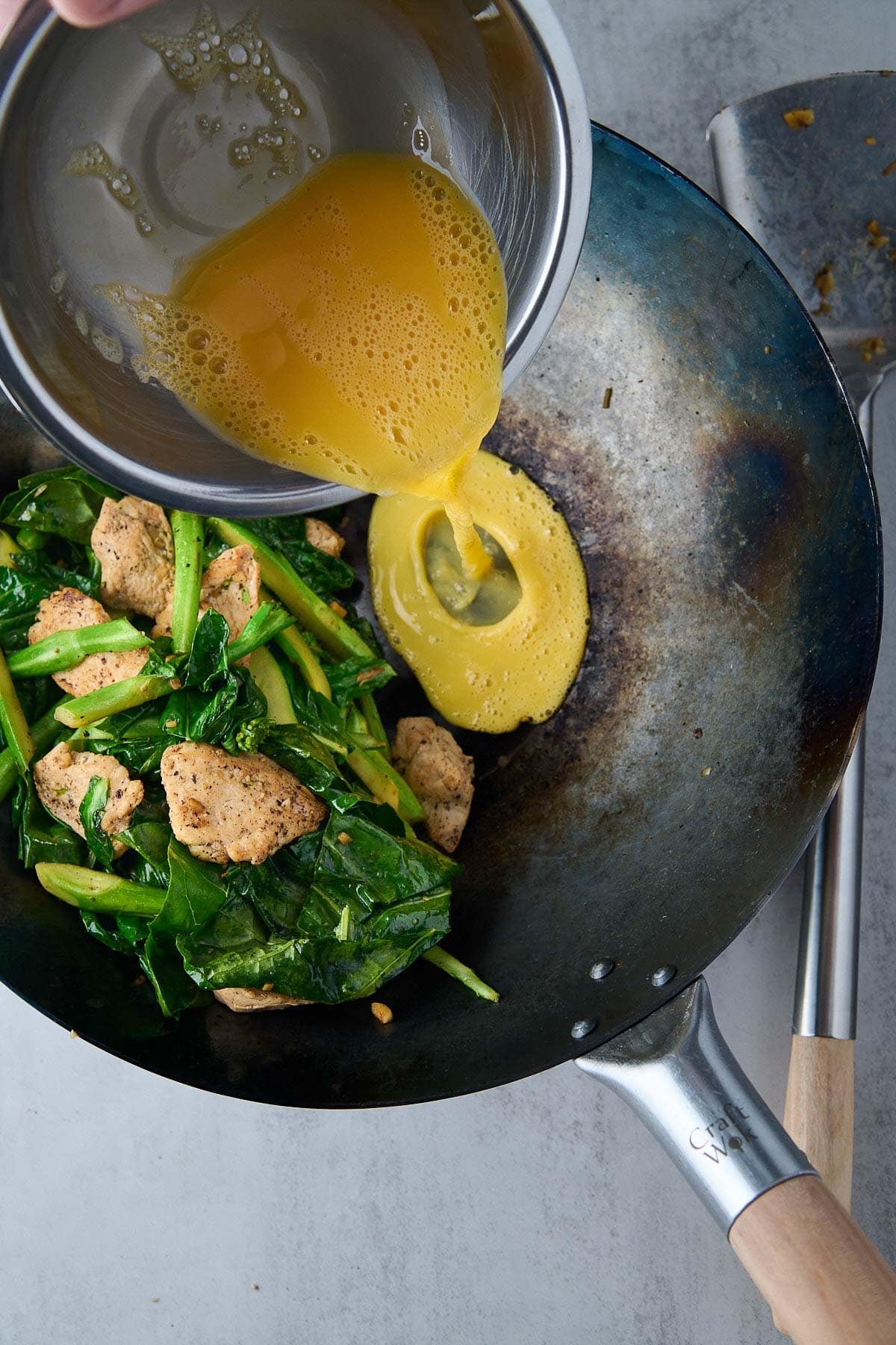 pouring whisked eggs in wok with chicken and Chinese broccoli.