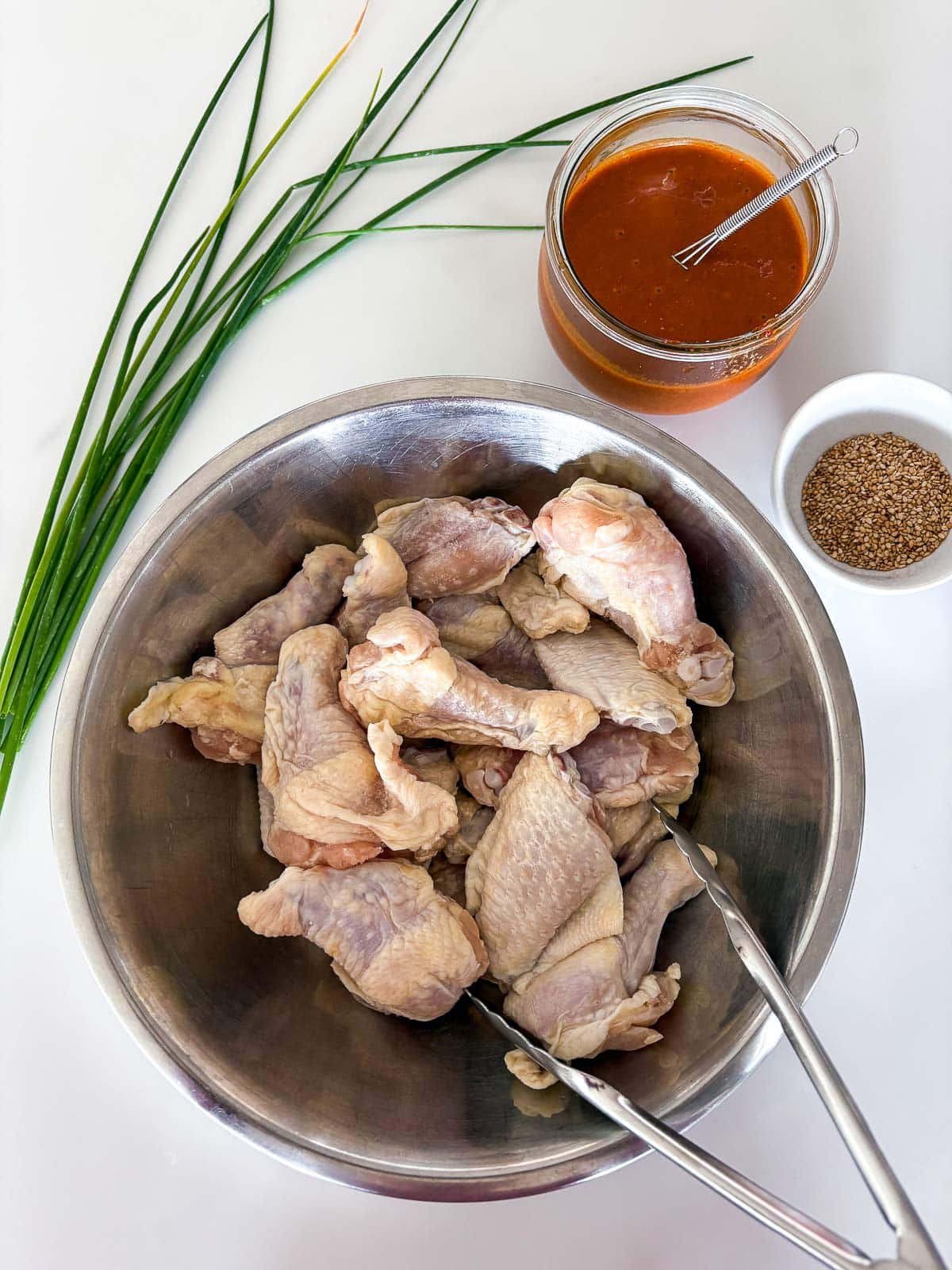 ingredients for sticky Asian chicken wings - chives, jar of marinade, bowl of toasted sesame seeds, metal mixing bowl with chicken wings and tongs. 