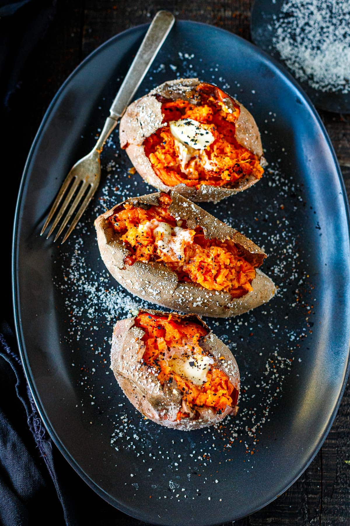 three baked potatoes topped with butter and chili flakes with salted skins on oval plate with fork. 