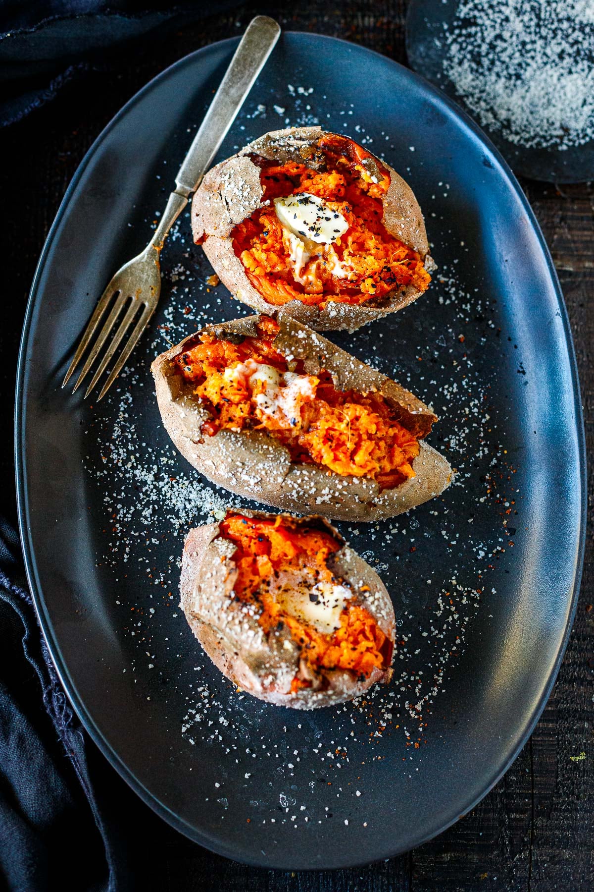 three baked sweet potatoes on a plate with a fork, topped with butter and salt.