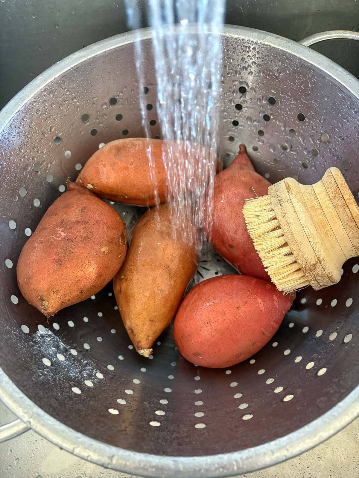colander with five sweet potatoes being rinsed with water with scrub brush. 