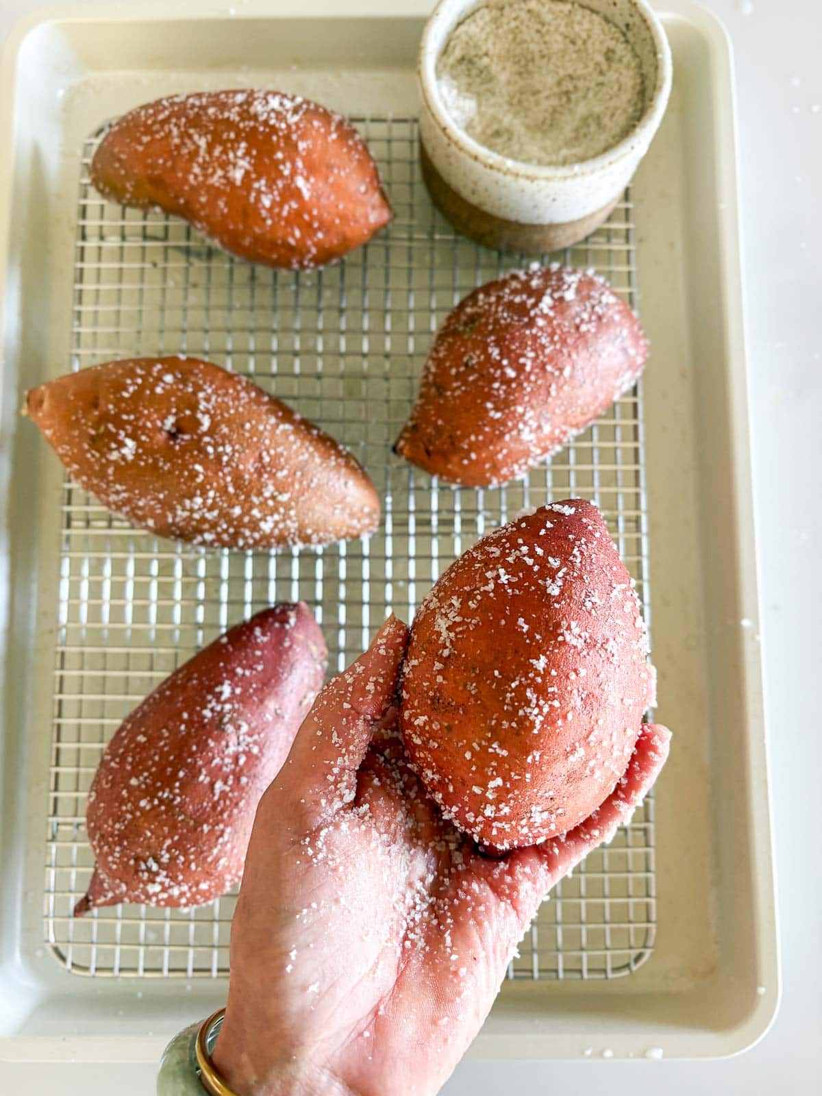 hand holding a salted sweet potato above a wire rack on sheet pan with four other sweet potatoes coated in salt next to salt dish. 