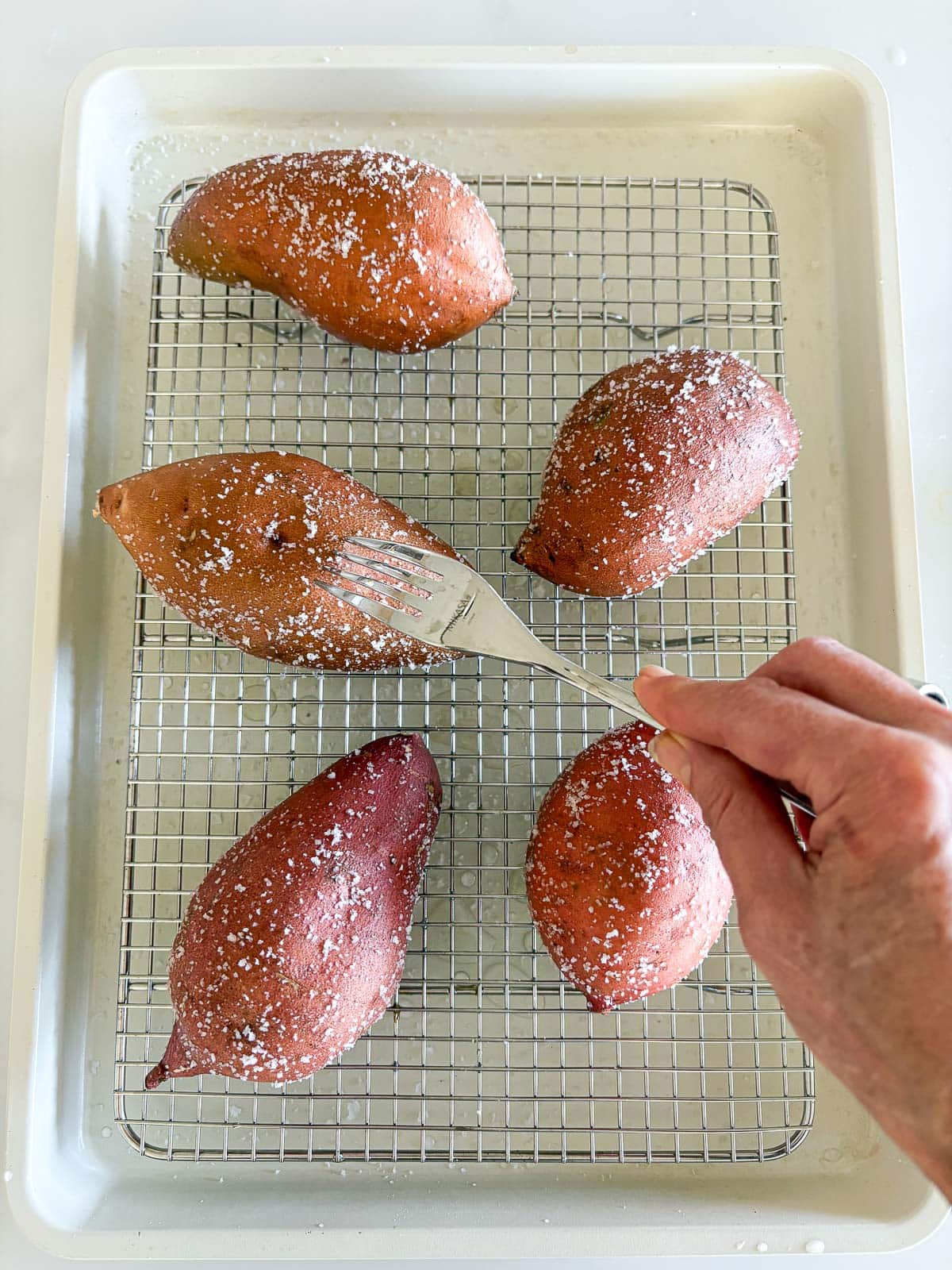 hand using fork to prick salted sweet potatoes on wire rack on baking sheet before baking. 
