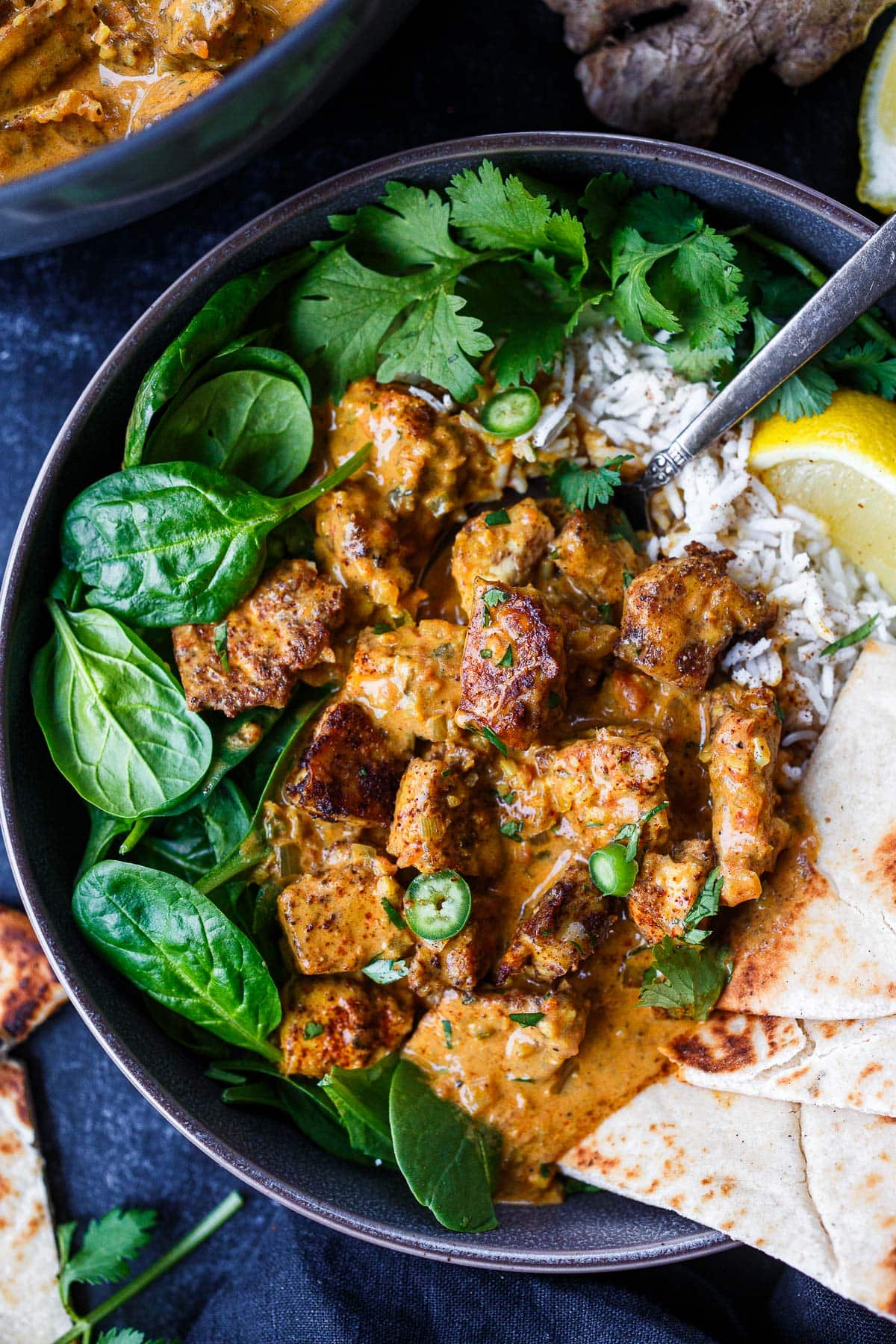 bowl of butter tofu on basmati rice and baby spinach with naan, lemon, sliced serrano chili, and cilantro.