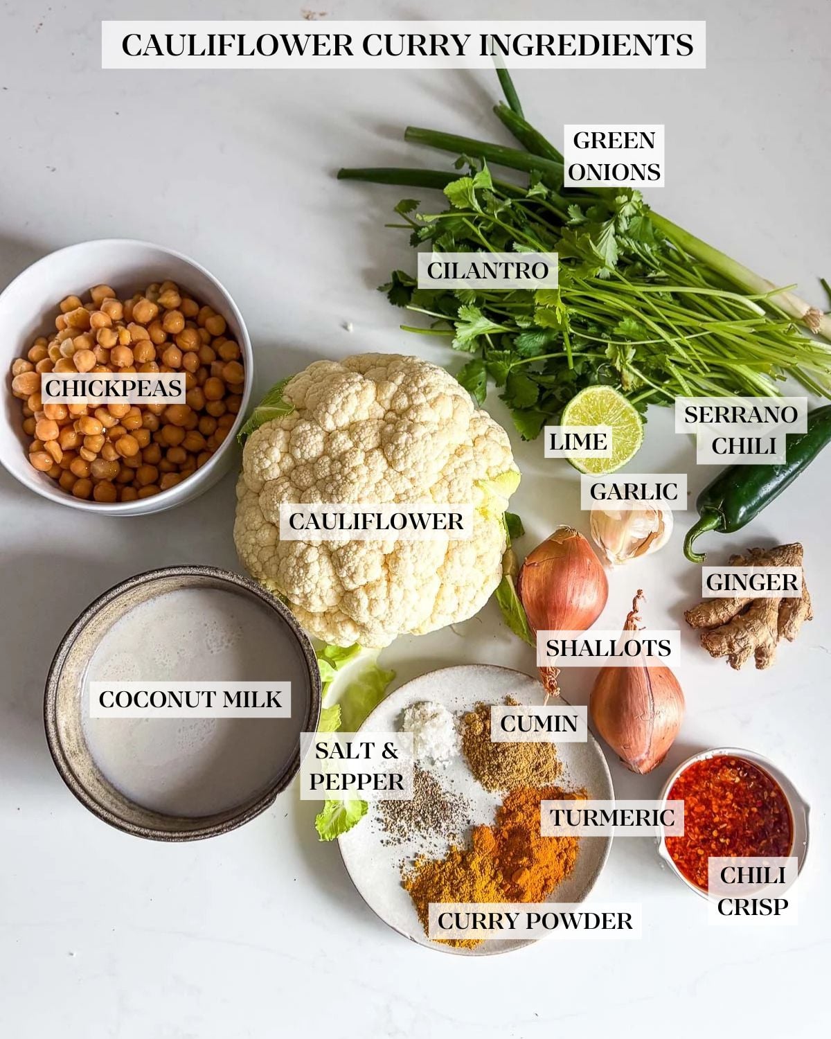 ingredients for cauliflower curry - green onions, cilantro, lime half, jalapeño, garlic, shallots, ginger, cauliflower head, chickpeas, coconut milk, small dish of spices, and small dish of chili crisp.
