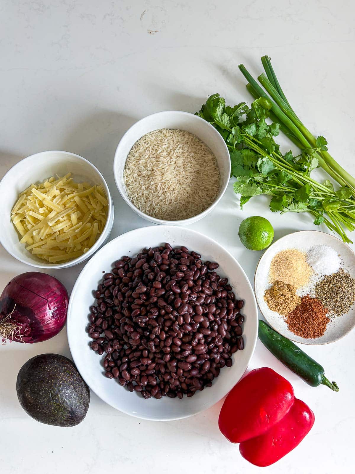 ingredients for cheesy rice and beans - fresh cilantro, rice, grated cheese, black beans, lime, spices, jalapeno, bell pepper, avocado, red onion.