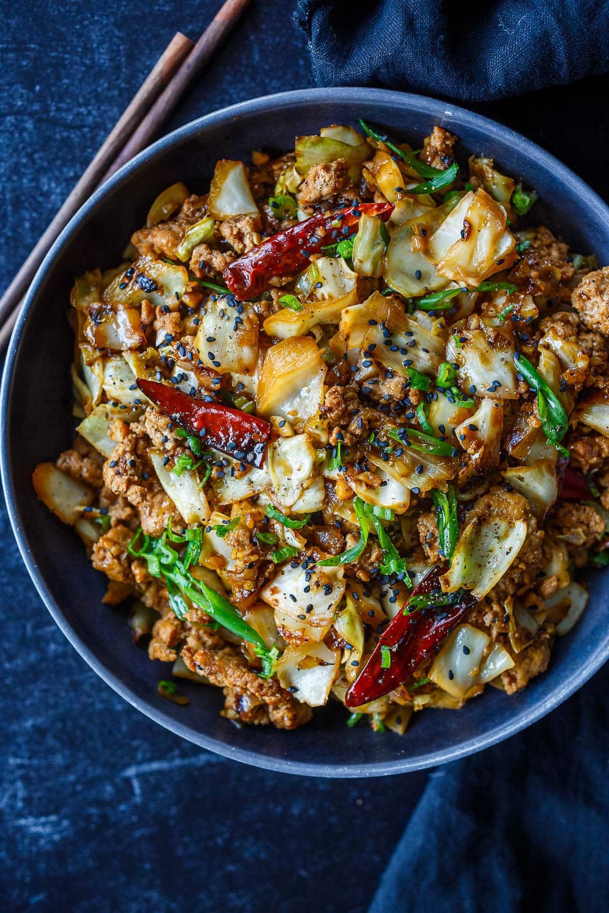 bowl of chicken and cabbage stir fry with dried red chilies, sesame seeds, and scallions with chopsticks on the side. 