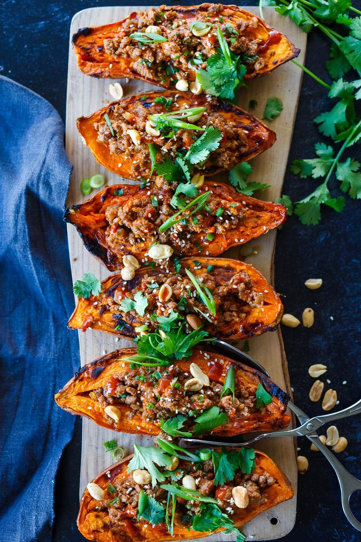 six gochujang ground turkey sweet potatoes on wood serving board stuffed with meat and topped with fresh herbs, sesame seeds, and peanuts.