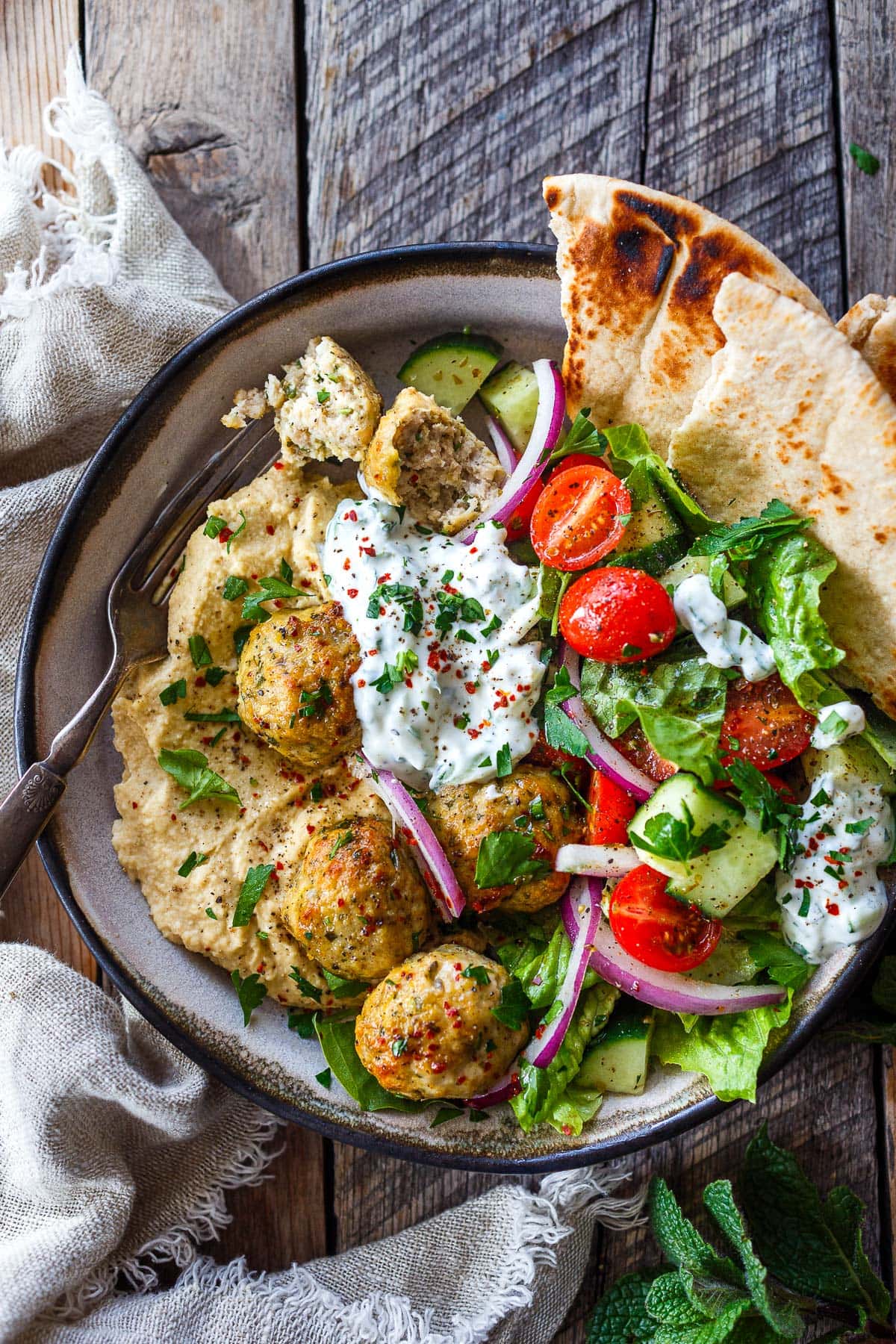 hummus bowl topped with Greek chicken meatballs, tzatziki, romaine, cherry tomatoes, red onion, cucumber, and parsley, with pita bread.