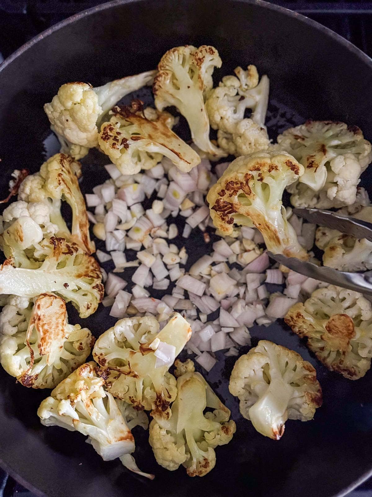 tongs flipping seared cauliflower florets with chopped shallots and garlic in center of pan.