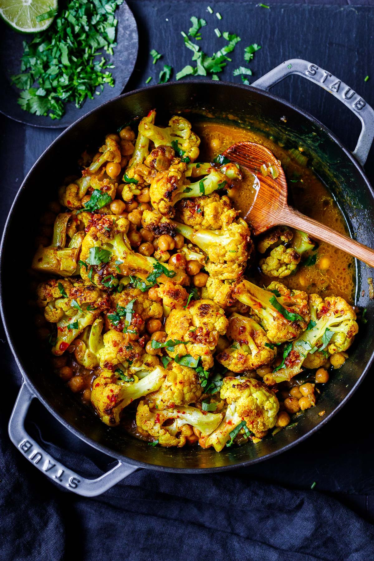 Dutch oven with cauliflower curry with chickpeas, fresh cilantro, and chili crisp with wood spoon.