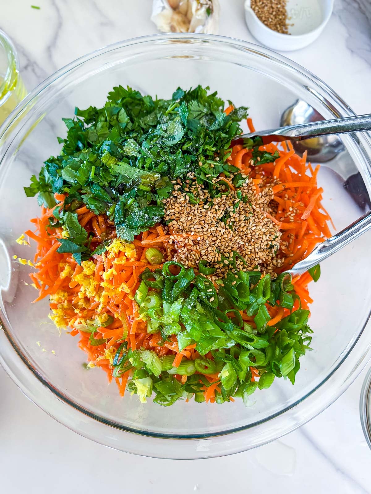 glass mixing bowl with all of the components to make asian carrot salad, including sesame seeds, cilantro, green onions, shredded carrots, ginger.