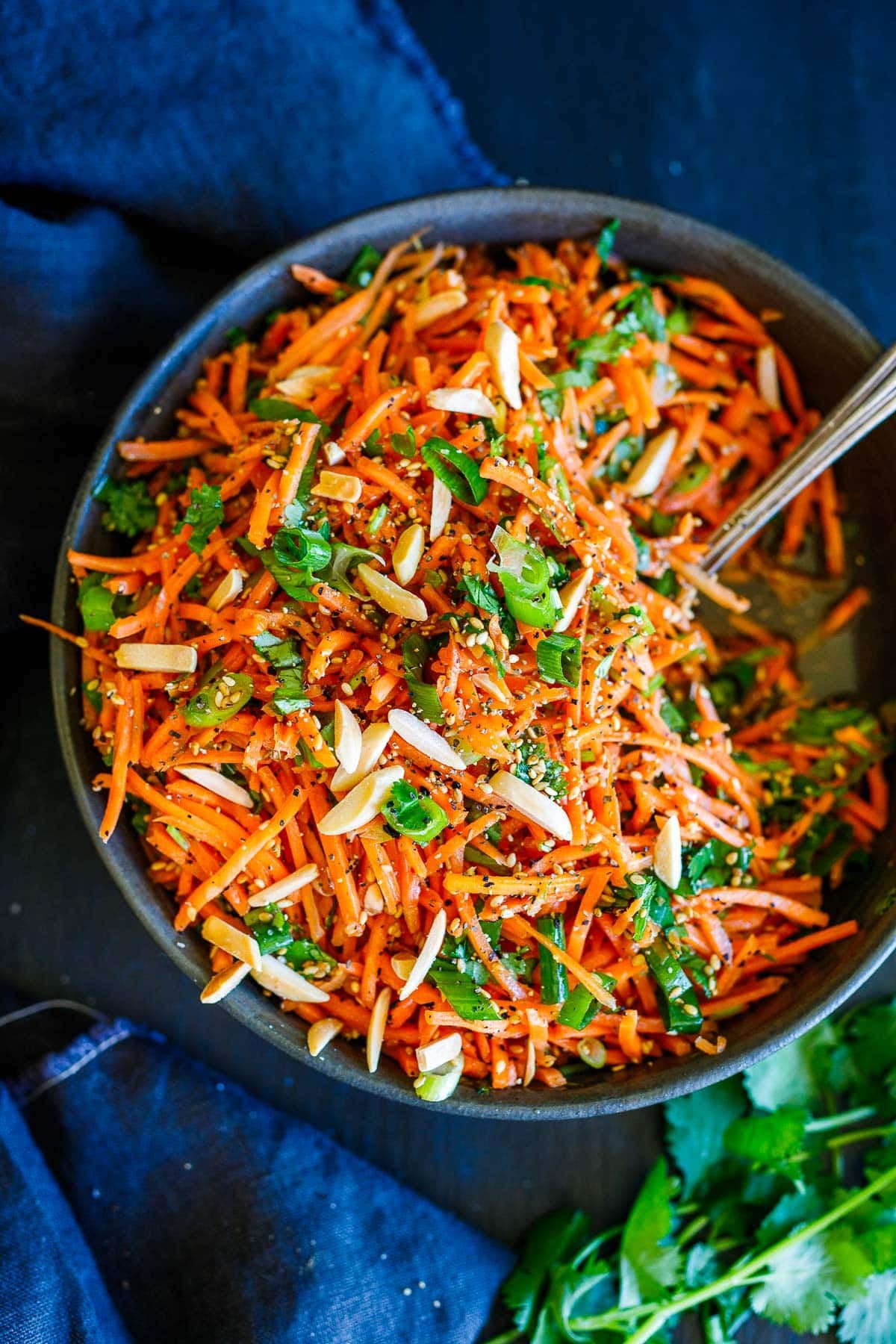 bowl of Asian carrot salad with green onions, toasted almonds, cilantro, and sesame seeds with fork lifting the salad up to reveal some of the dressing in the bowl.