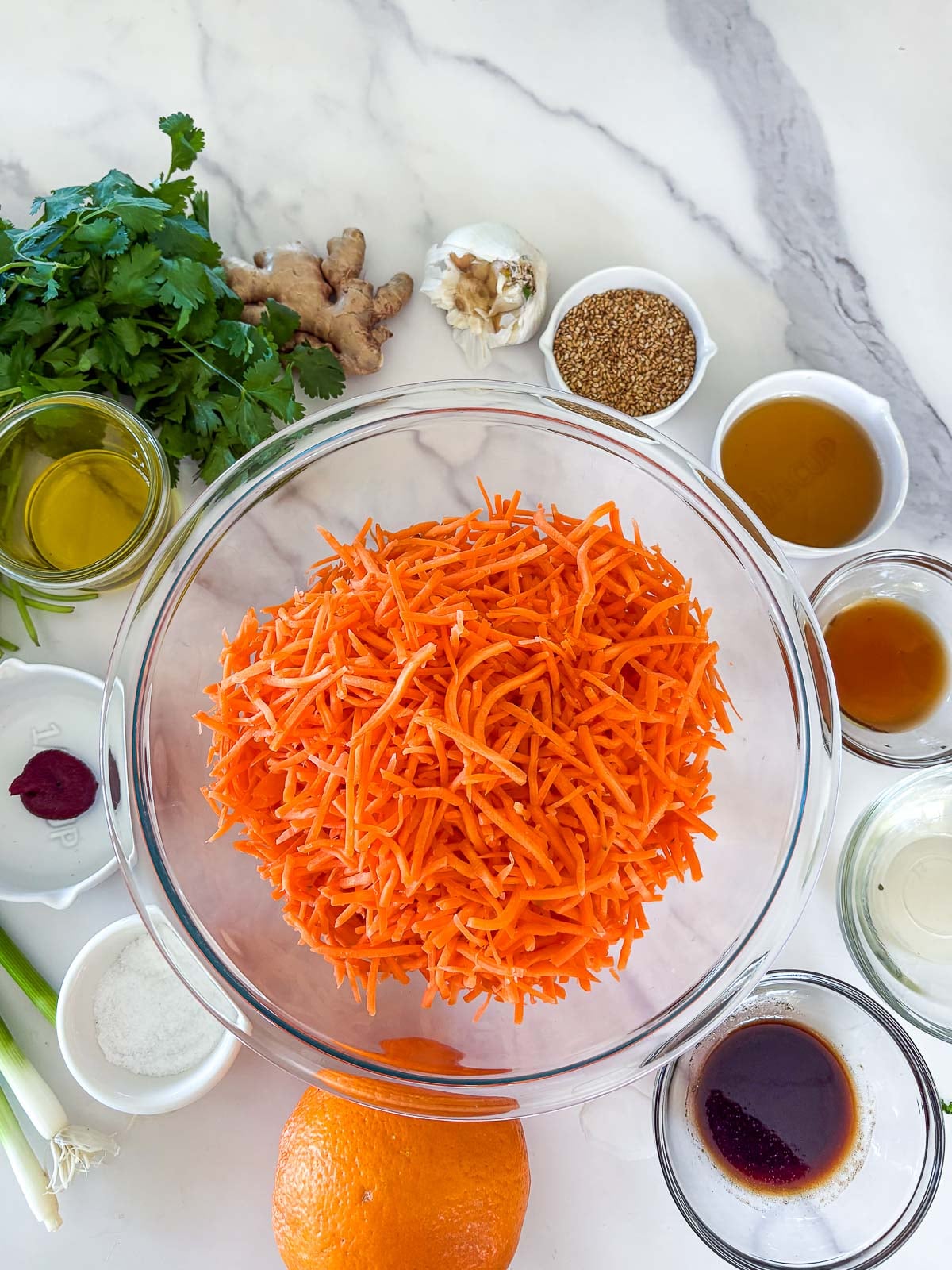 bowl of shredded carrots surrounded by remaining ingredients to make asian carrot salad, including cilantro, ginger, garlic, sesame seeds, orange, and green onions, with small bowls of sauces.