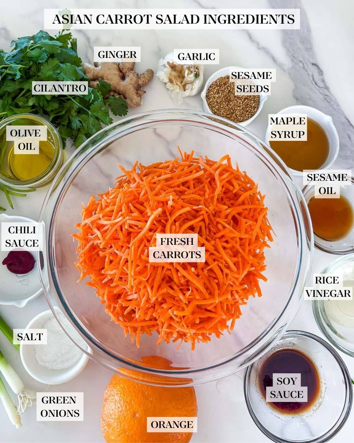 bowl of shredded carrots surrounded by remaining ingredients to make asian carrot salad, including cilantro, ginger, garlic, sesame seeds, orange, and green onions, with small bowls of sauces. 