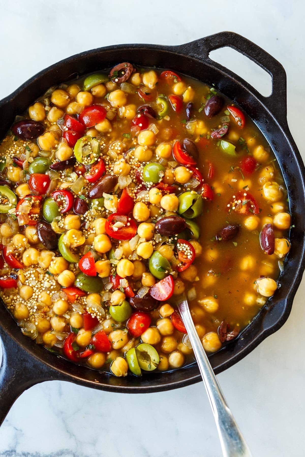 assembling Mediterranean chickpea skillet in cast iron skillet with quinoa, chickpeas, olives, tomatoes, and broth. 