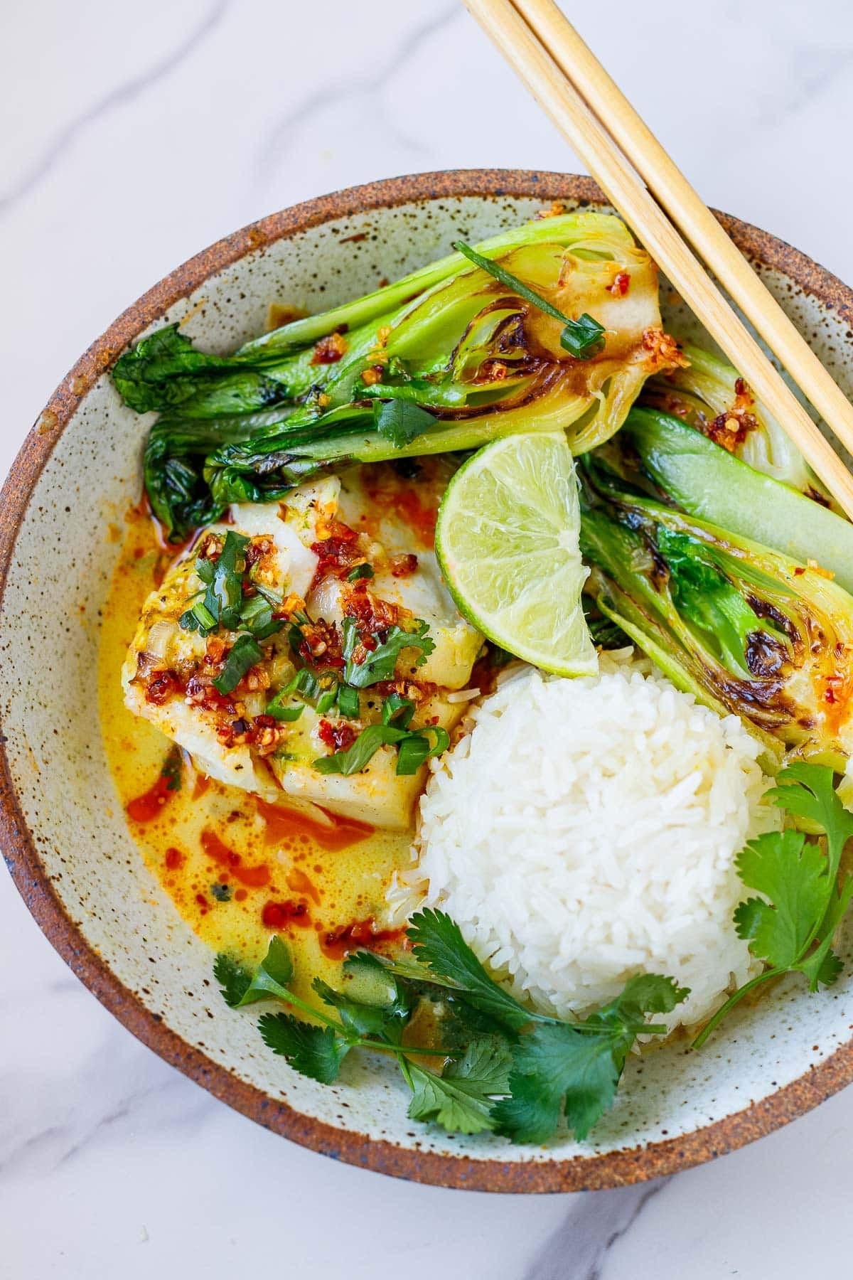 coconut cod recipe in bowl of coconut broth with jasmine rice, seared bok choy, lime, and chili crisp, with fresh cilantro and chopsticks.