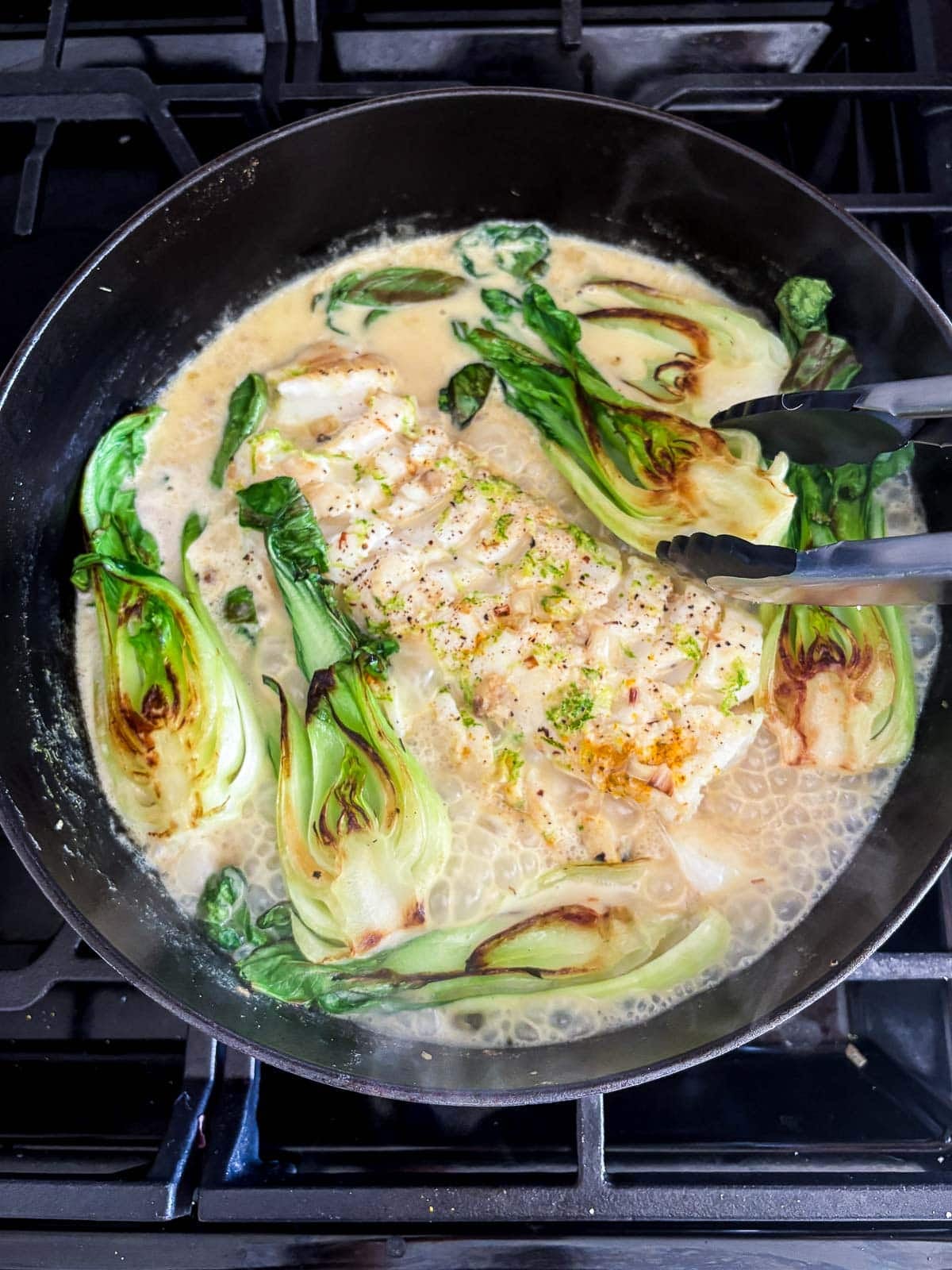 cod recipe in pot of coconut broth with seared bok choy and lime zest with tongs.