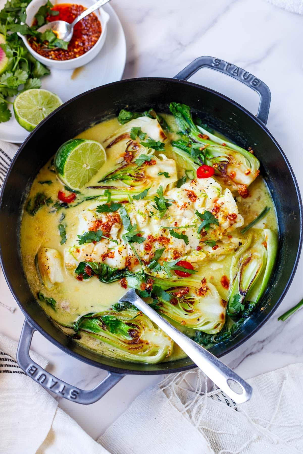 coconut cod recipe in pot of creamy broth with seared bok choy, red chilis, chili crisp, lime half, and fresh herbs.