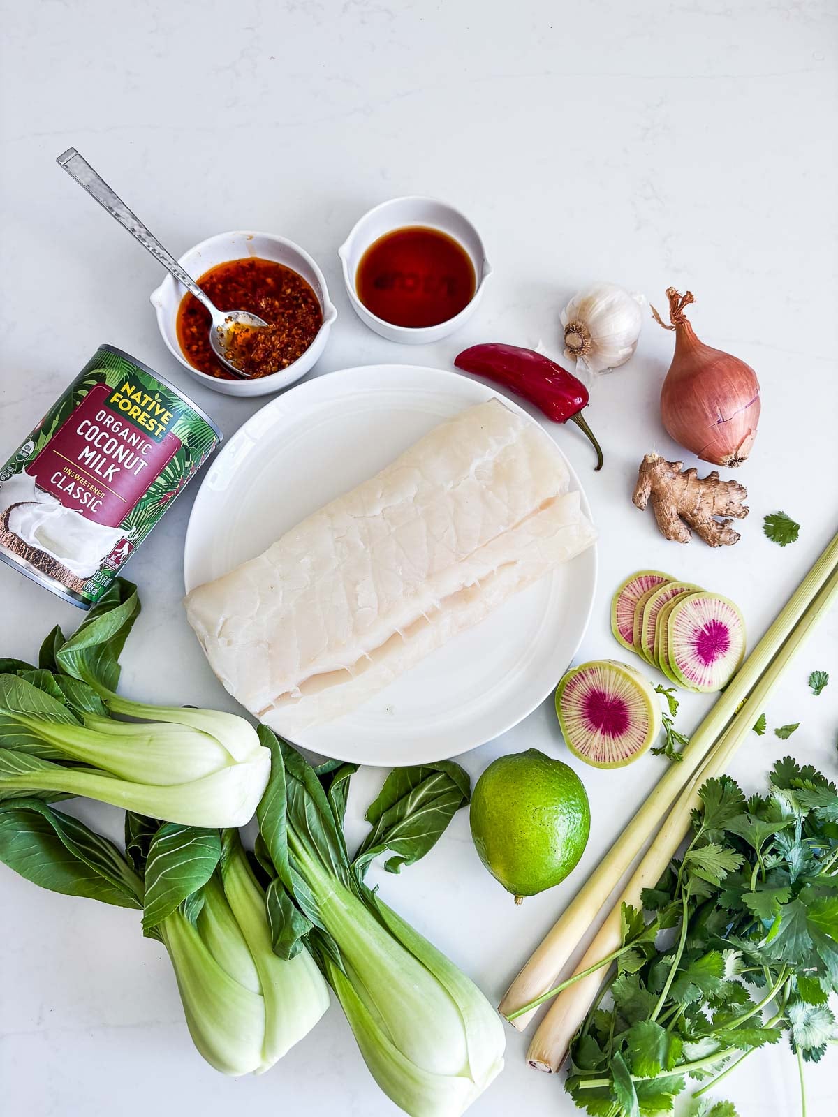 coconut cod recipe ingredients - uncooked cod on plate, coconut milk, chili crisp, soy sauce, red chili, garlic, shallot, ginger, sliced watermelon radish, lime, lemongrass, fresh cilantro, baby bok choy.