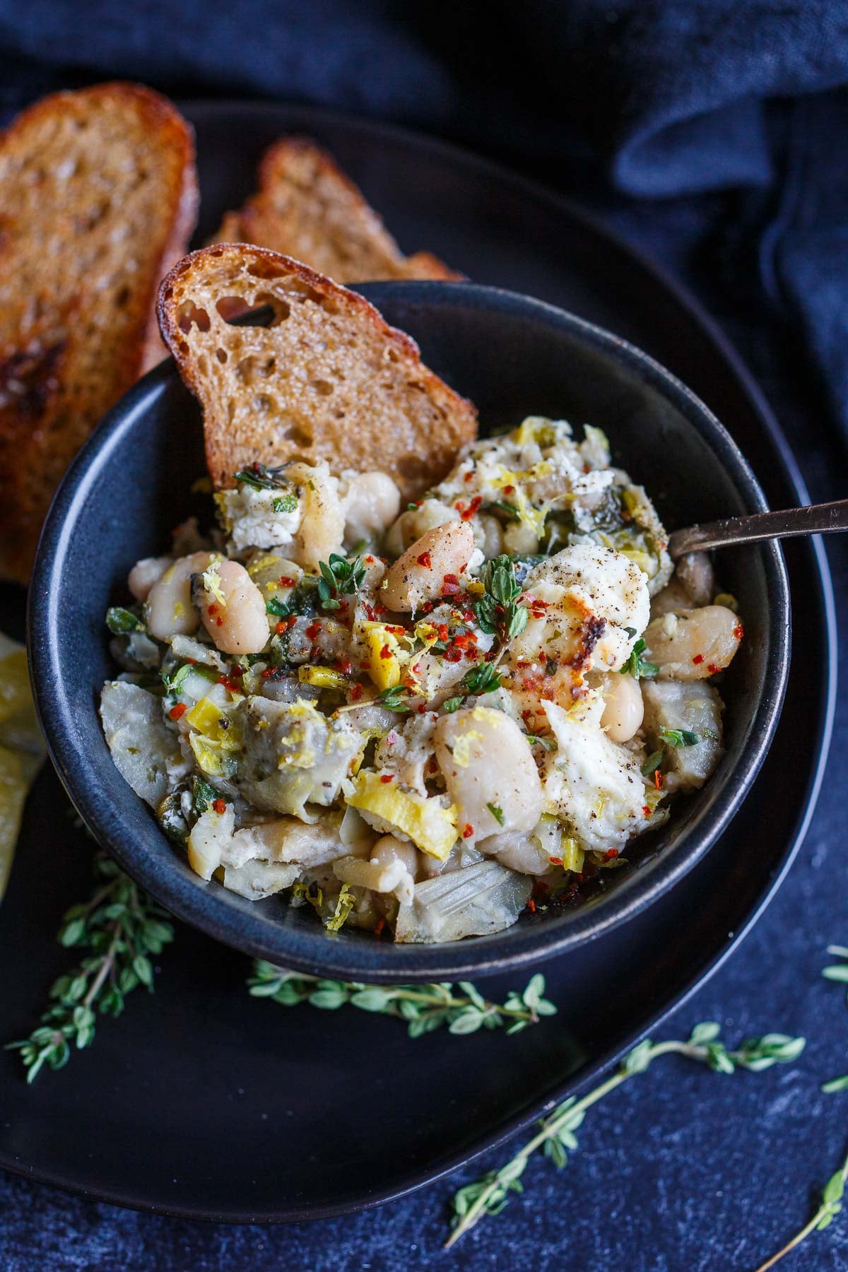 bowl of white bean artichoke skillet with lemon zest, herbs, and red pepper flakes with toasted sourdough.