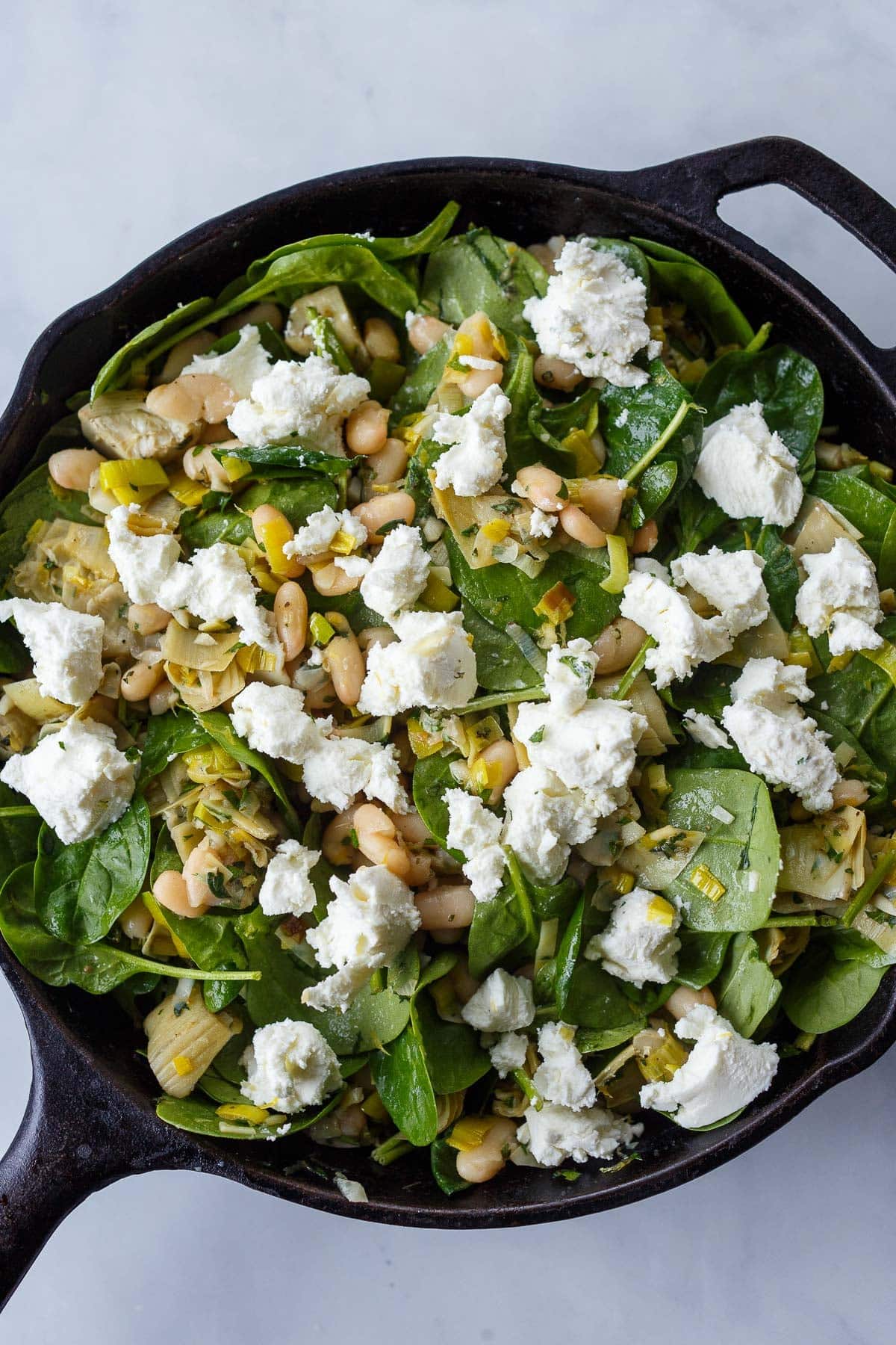 assembled white bean artichoke skillet before baking - fresh goat cheese on top of spinach, white beans, artichokes, and sautéed leeks.