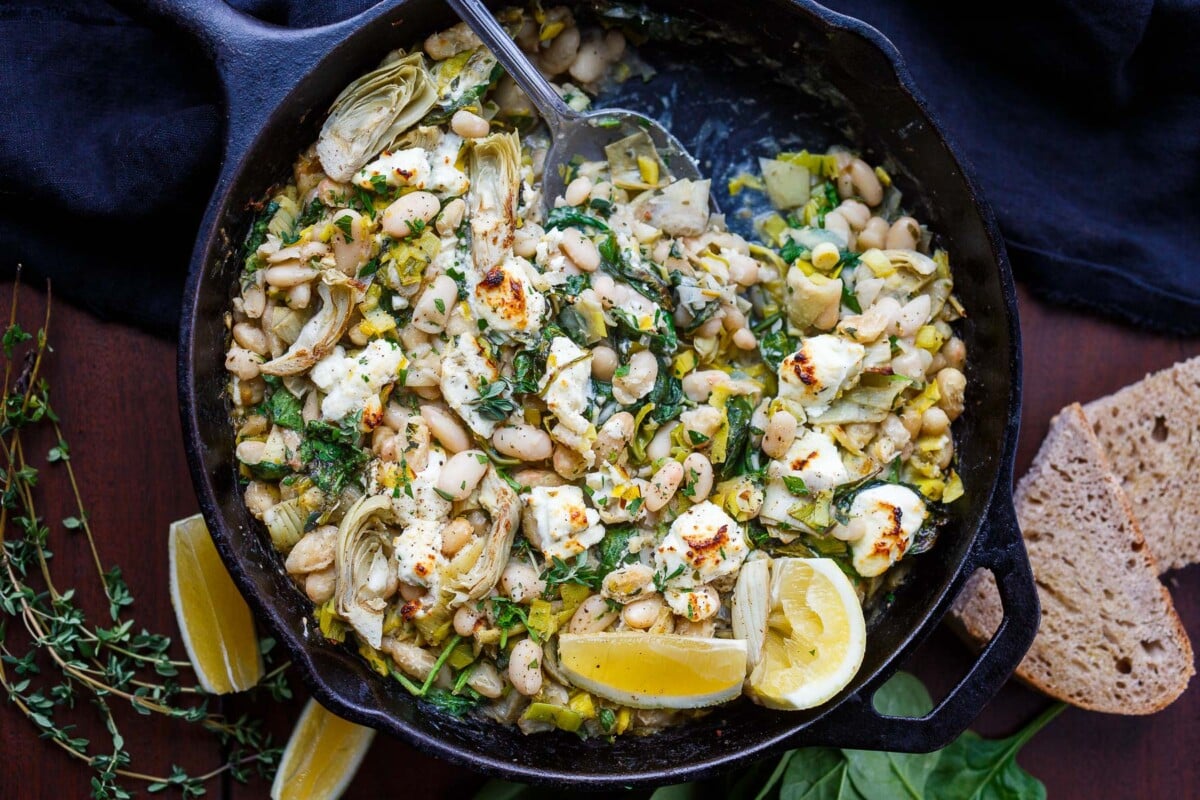 white bean skillet dinner with artichokes, leeks, goat cheese, and herbs with lemon wedges and spoon for serving.