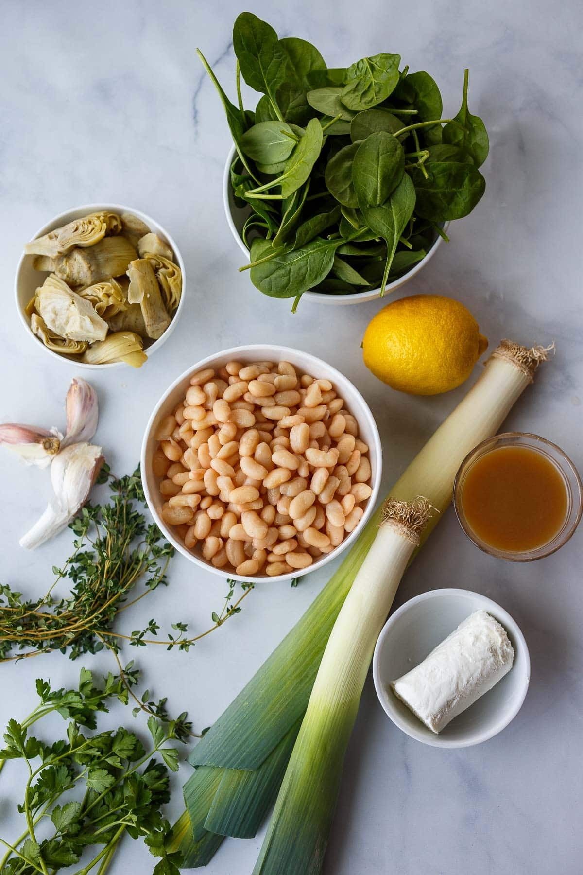 ingredients for white bean artichoke skillet neatly arranged - fresh spinach, artichoke hearts, lemon, bowl of white beans, garlic cloves, fresh herbs, leeks, vegetable broth, and goat cheese.