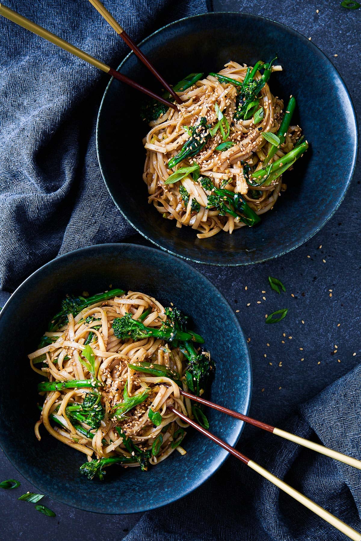 two bowls with garlic noodles with broccolini, scallions, sesame seeds, and chopsticks. 