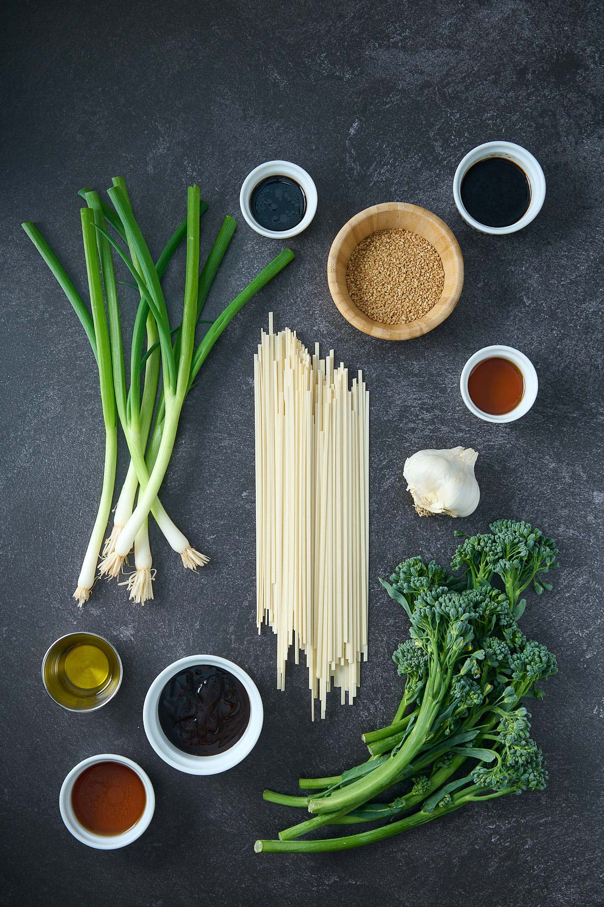 ingredients for garlic noodles - scallions, sesame seeds, noodles, garlic, broccolini, and various sauces in small bowls. 