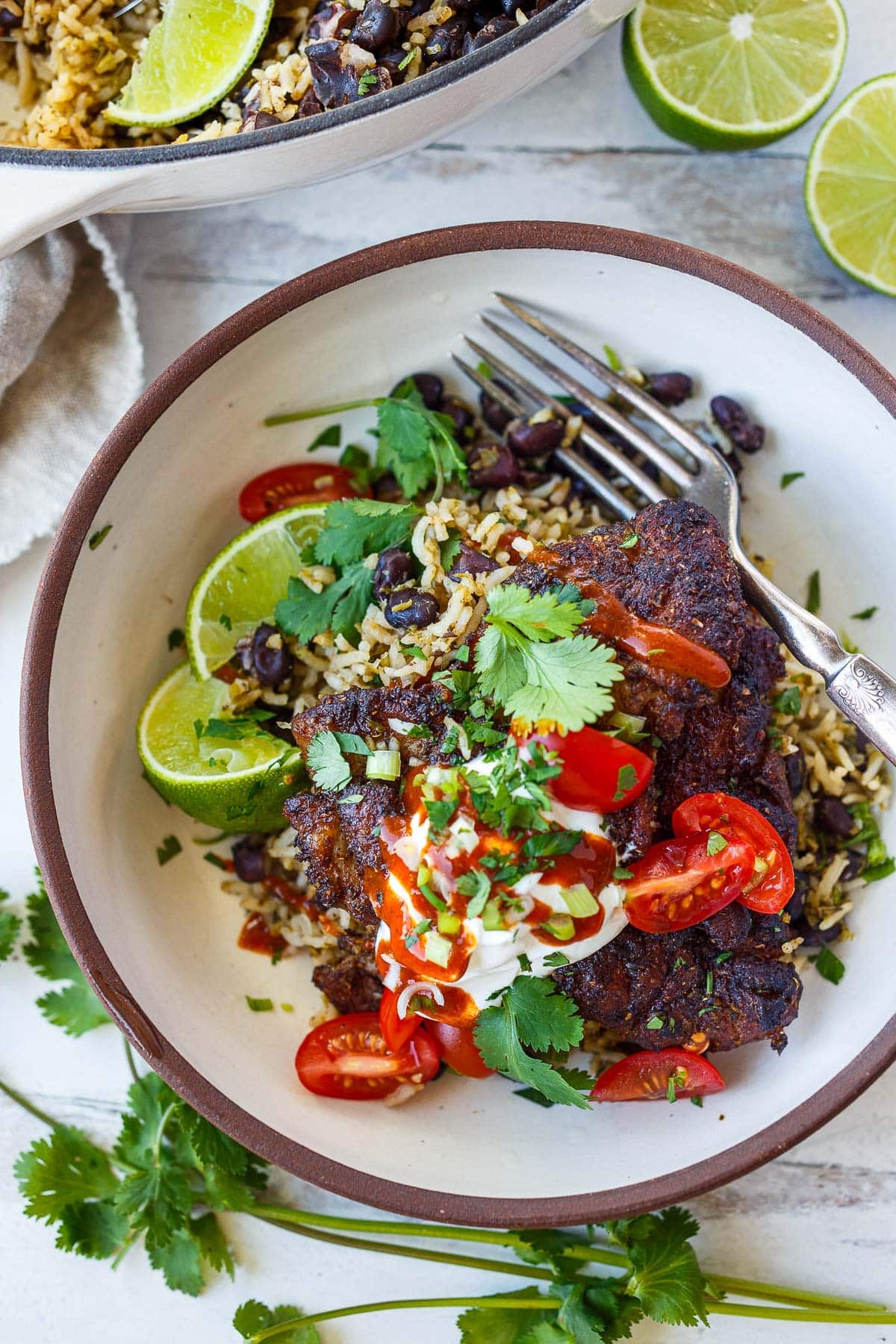 Mexican chicken, rice, and beans in bowl with lime wedges, cilantro, green onions, tomatoes, sour cream, hot sauce, and a fork.