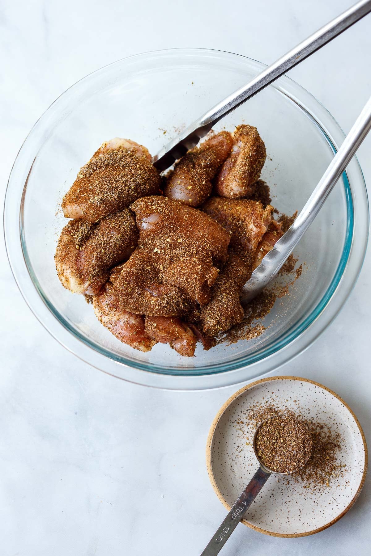 glass mixing bowl with dry rubbed chicken thighs and tongs. 