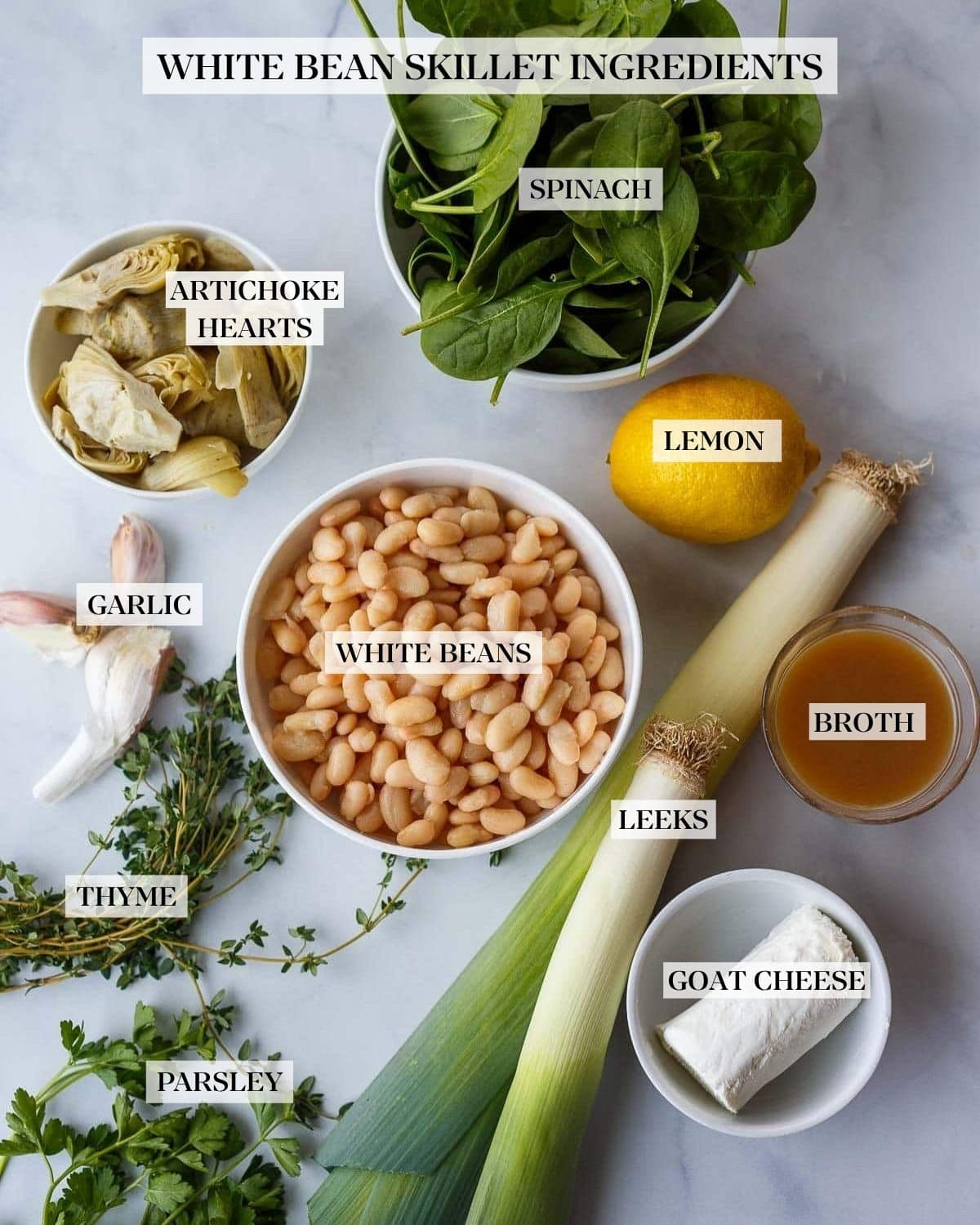 ingredients for white bean artichoke skillet neatly arranged - fresh spinach, artichoke hearts, lemon, bowl of white beans, garlic cloves, fresh herbs, leeks, vegetable broth, and goat cheese.