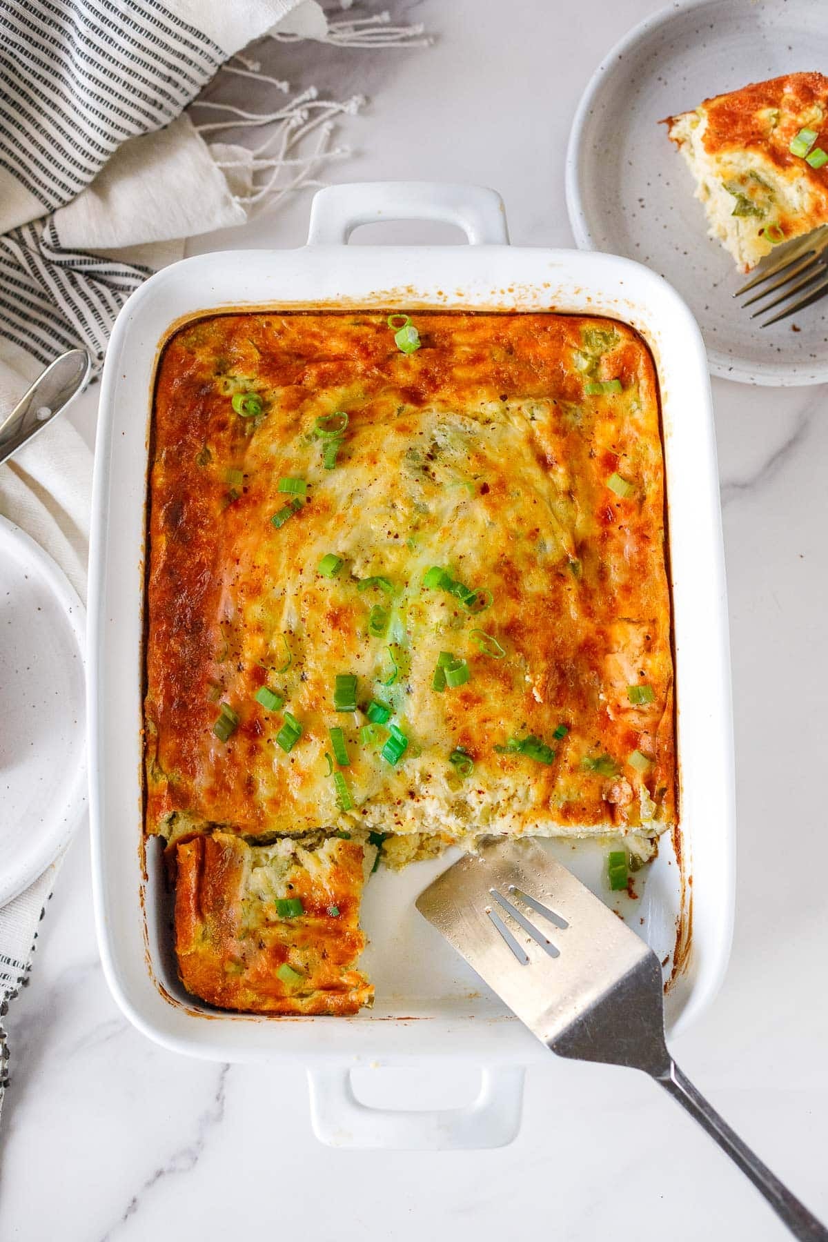 breakfast casserole with golden cheesy top and garnish of green onions, with two squares removed and one square cut, with metal spatula rest in the pan. 