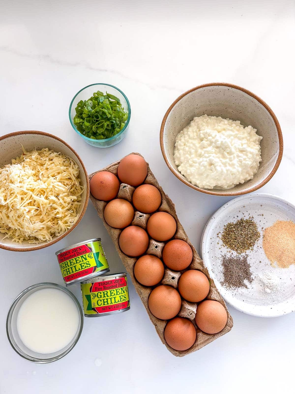 ingredients for easy breakfast casserole neatly arranged in bowls - green onions, cottage cheese, spices, eggs, two small cans of green chiles, bowl of milk, and grated cheese. 