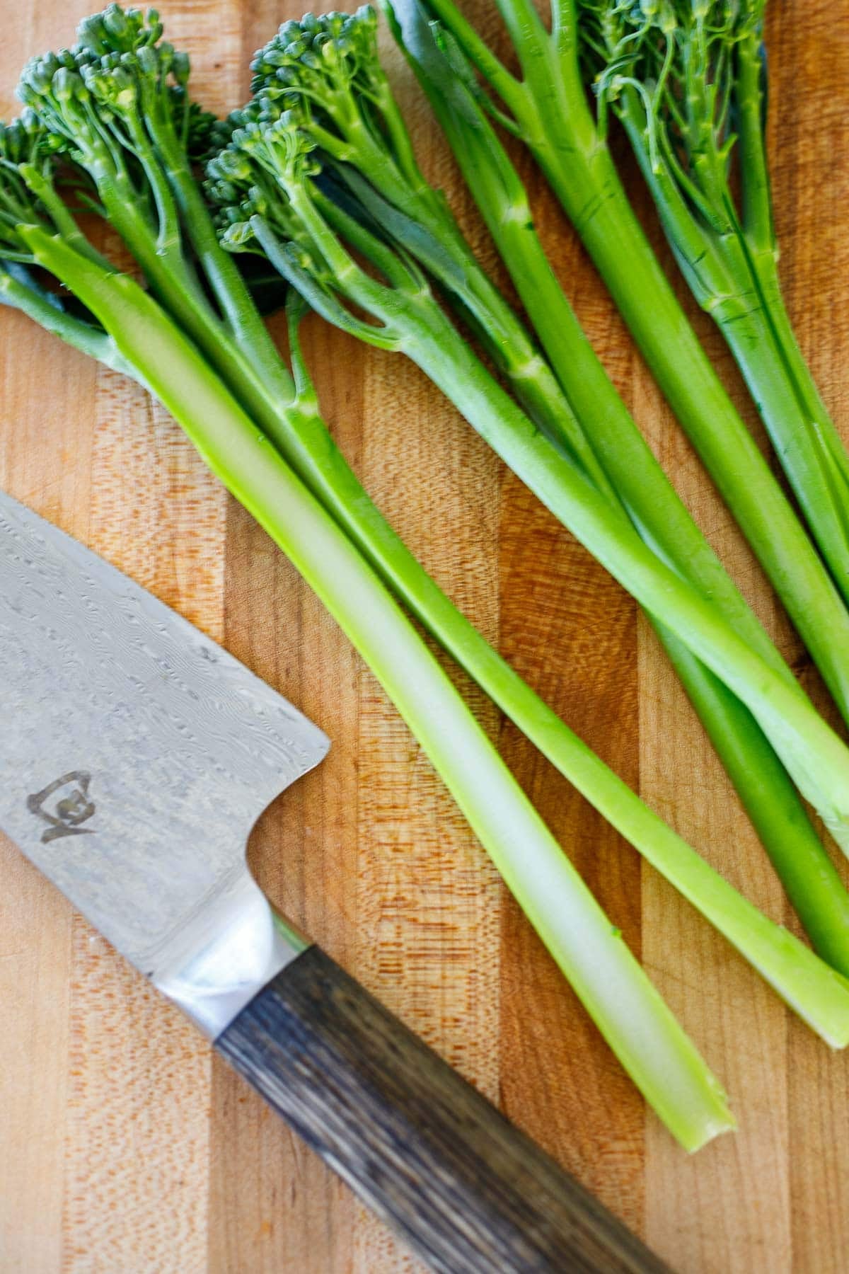 broccolini sliced in half lengthwise on wood cutting board with sharp knife.