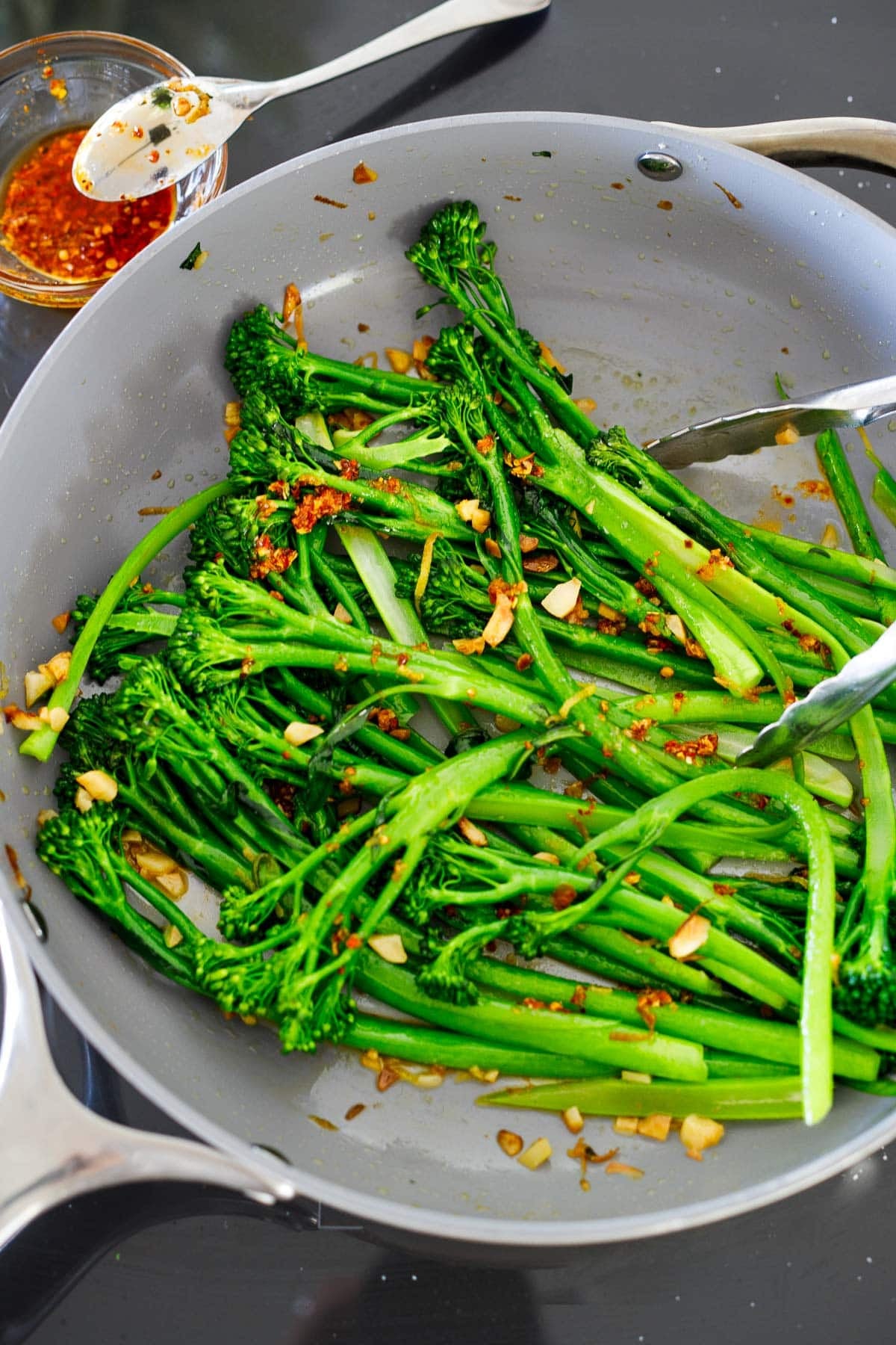 tender-crisp broccolini in sauté pan with garlic and chili crisp with tongs.
