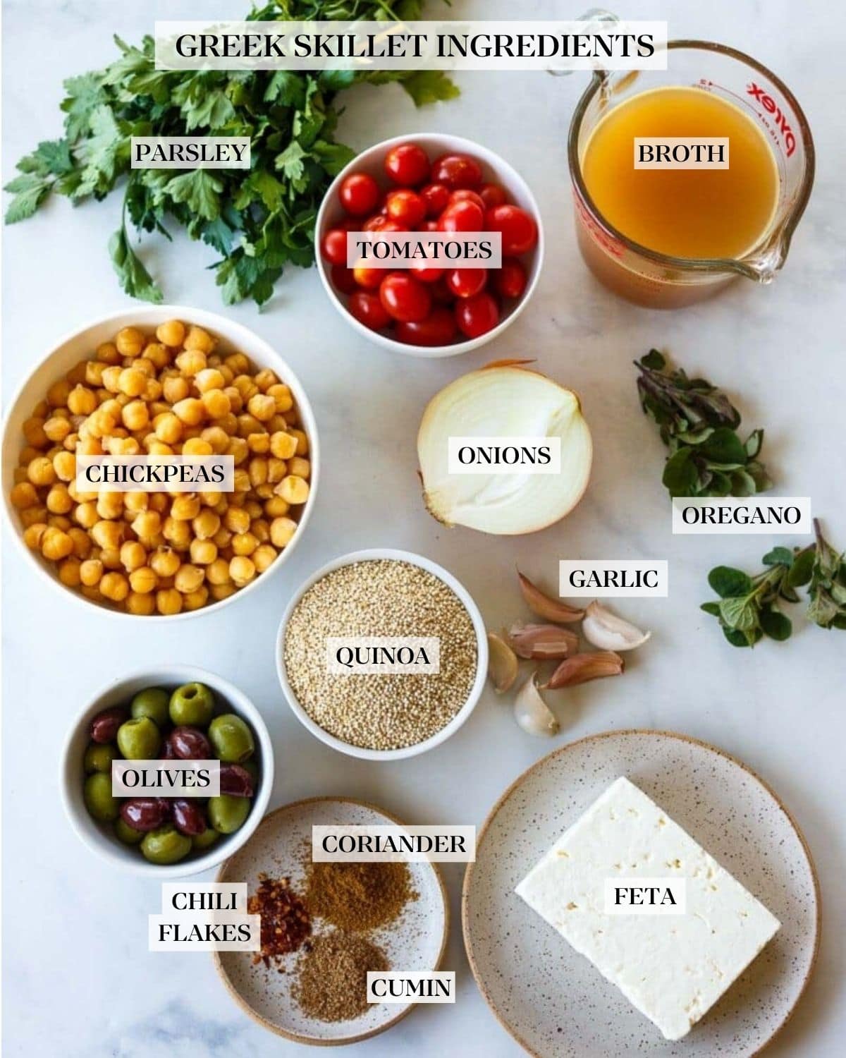 Chickpea meal ingredients arranged on countertop for Mediterranean chickpea skillet - parsley, broth, cherry tomatoes, chickpeas, onion, herbs, garlic cloves, quinoa, olives, spices, and feta block. 
