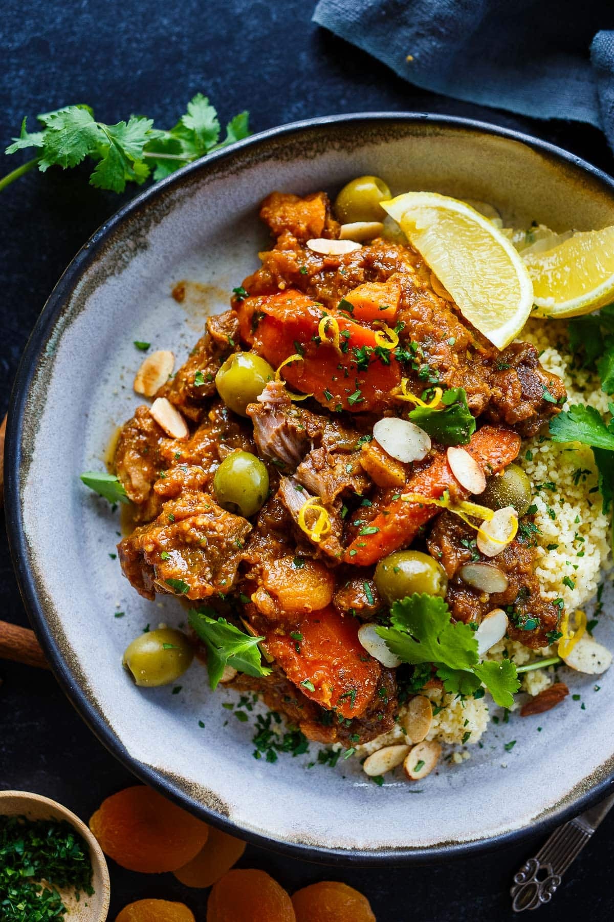 Moroccan lamb tagine on couscous with sliced almonds, fresh herbs, lemon zest, green olives, stewed carrots, and lemon wedges.