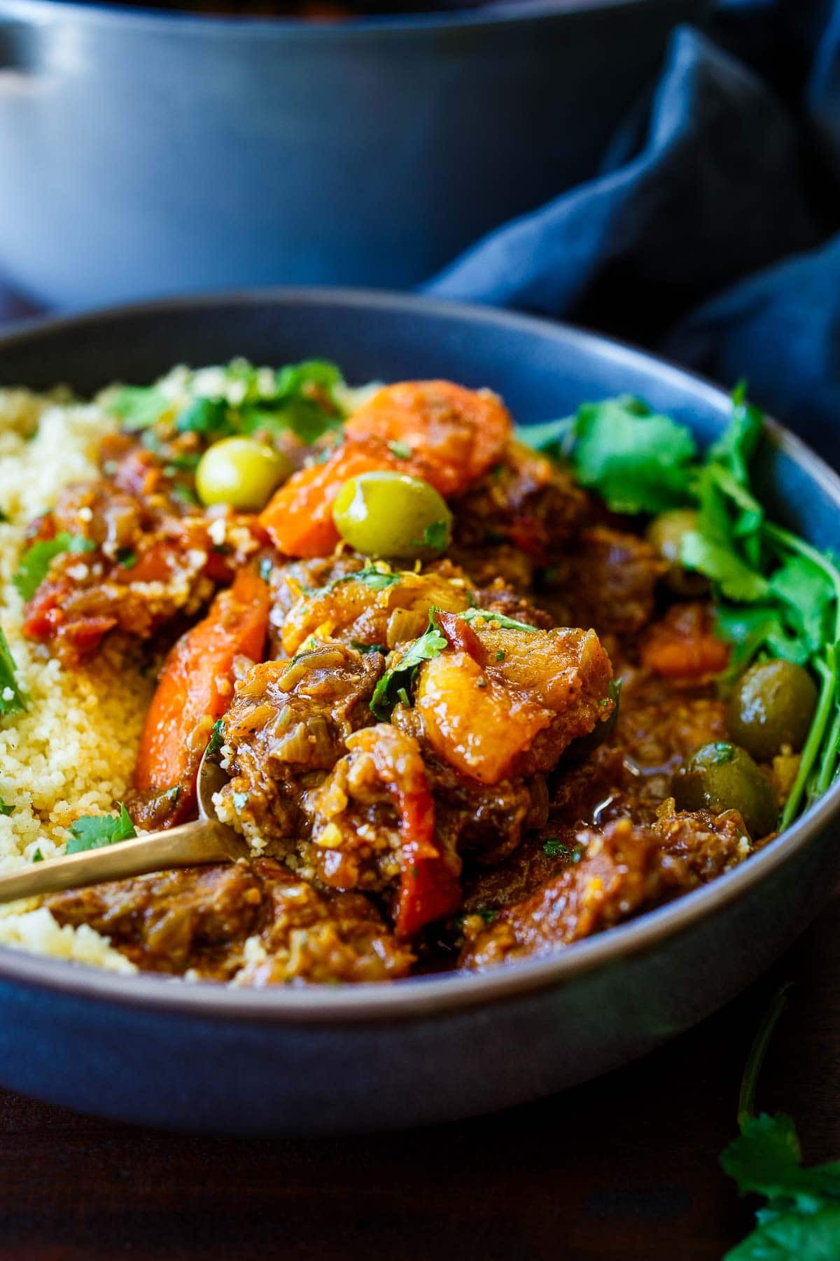 spoon scooping up lamb tagine from bowl with couscous, cilantro, and green olives.
