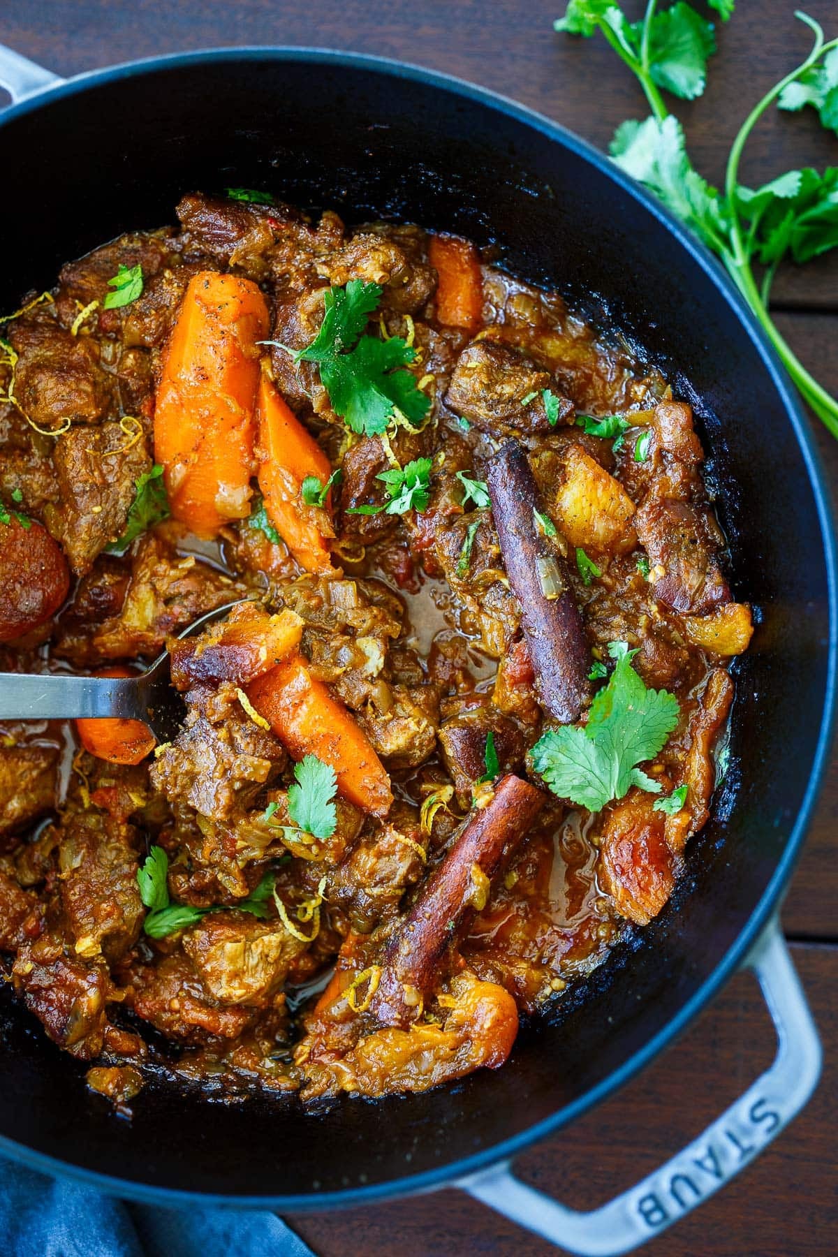 Moroccan lamb tagine in pot with carrots, dried apricots, cinnamon sticks, fresh cilantro, lemon zest, and a spoon.