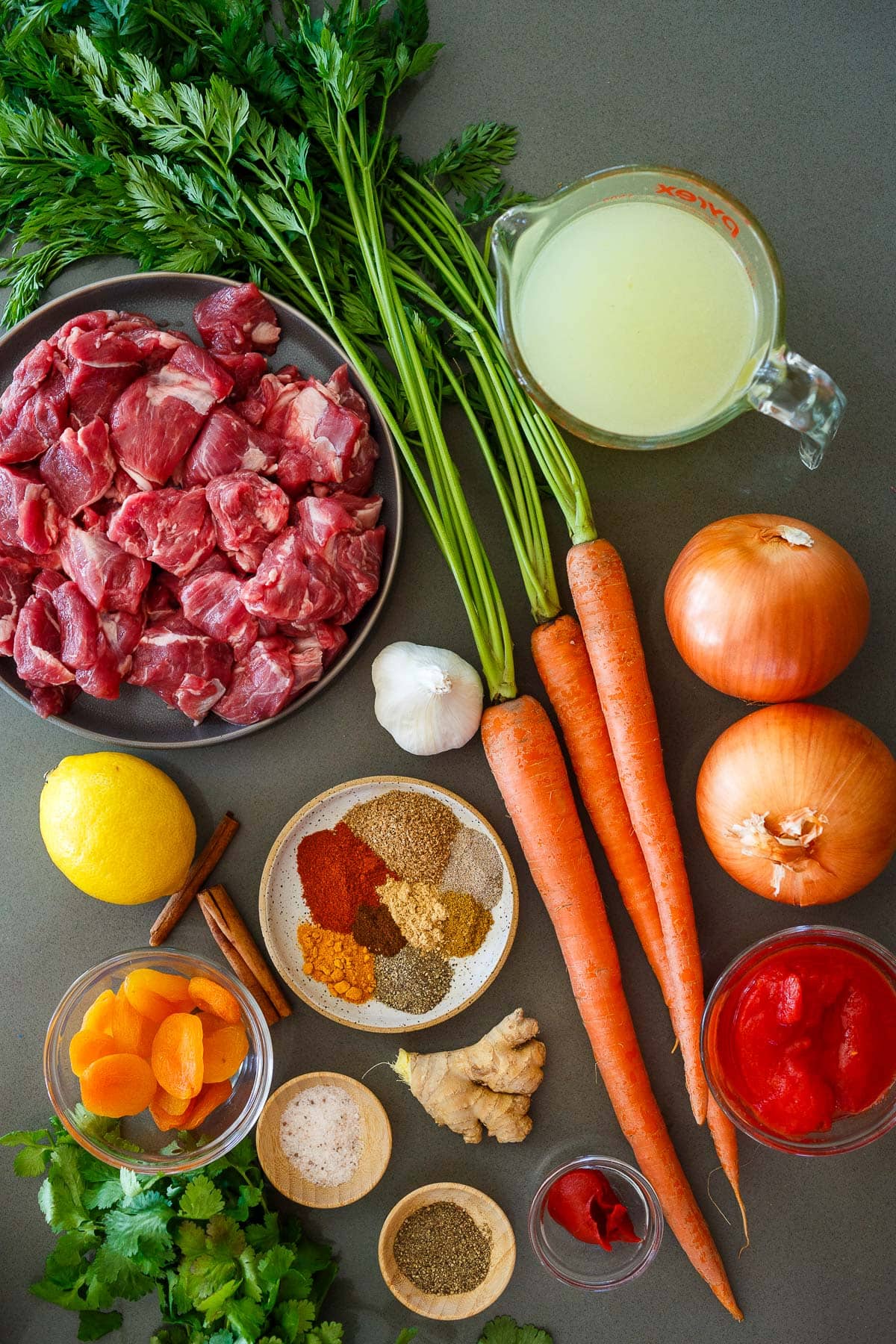 lamb tagine ingredients with plate of lamb shoulder, measuring cup of broth, two onions, three carrots, garlic bulb, small bowls of crushed tomatoes and tomato paste, plate of spices, fresh ginger, lemon, small bowl of dried apricots, two small bowls of salt and pepper, and fresh cilantro.